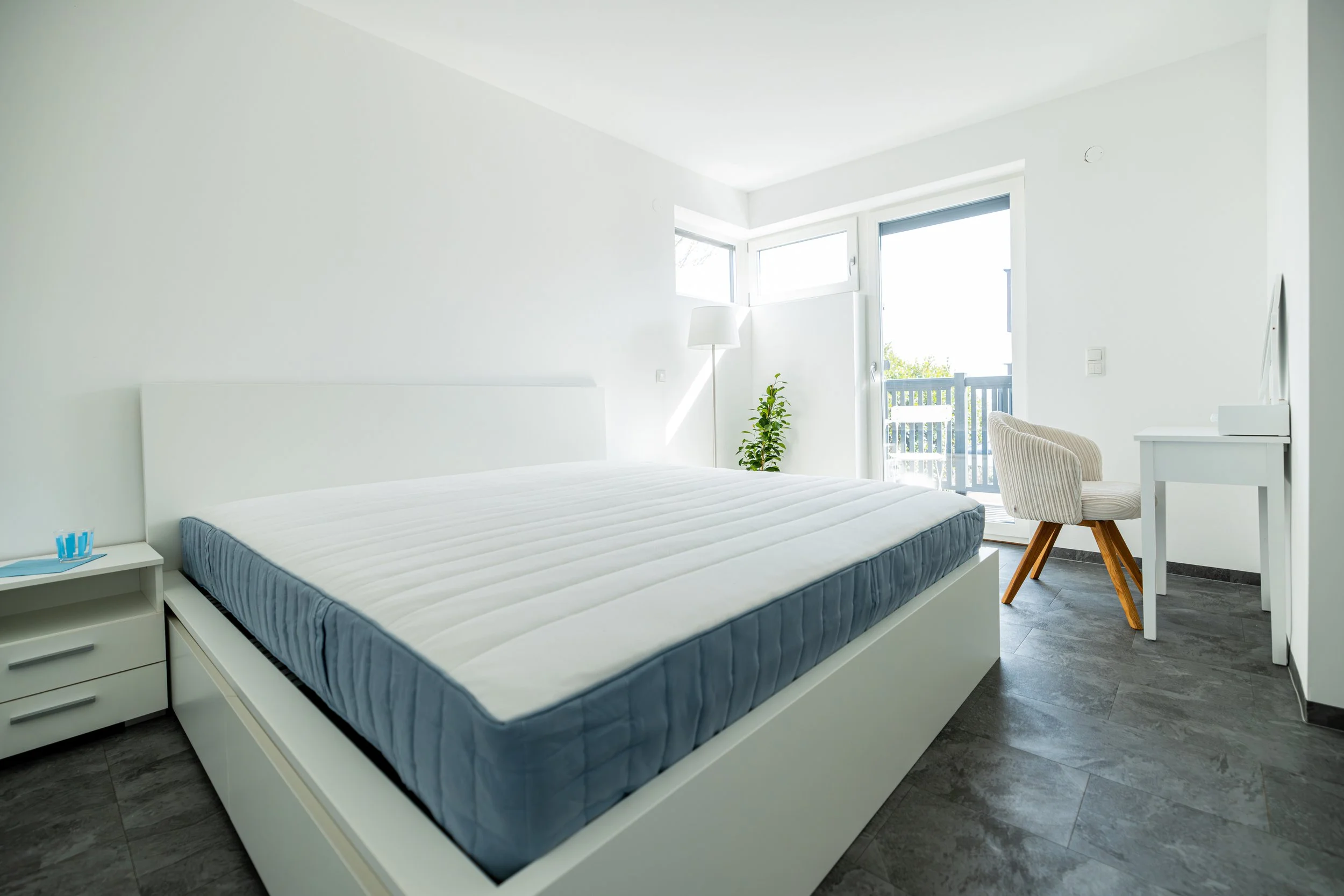 Schlafzimmer mit weißem Bett, Nachttisch, Stehlampe, Stuhl, Schreibtisch und Balkon mit Aussicht, helle und minimalistische Einrichtung.