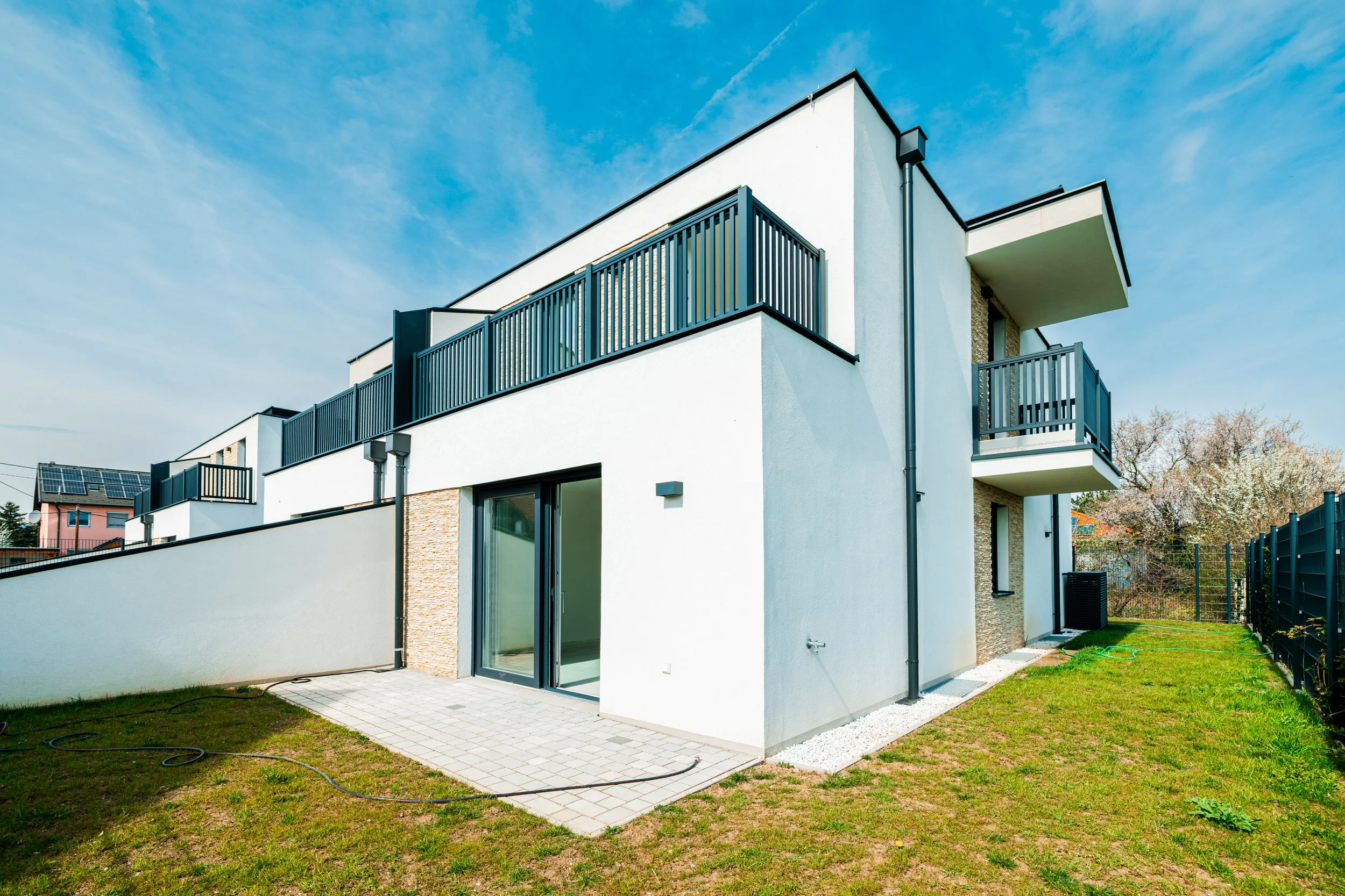 Neues modernes Wohnhaus mit weißen Wänden, mehreren Balkonen mit schwarzen Geländern, großer Terrasse mit Pflastersteinboden, grünem Garten und blauem Himmel.