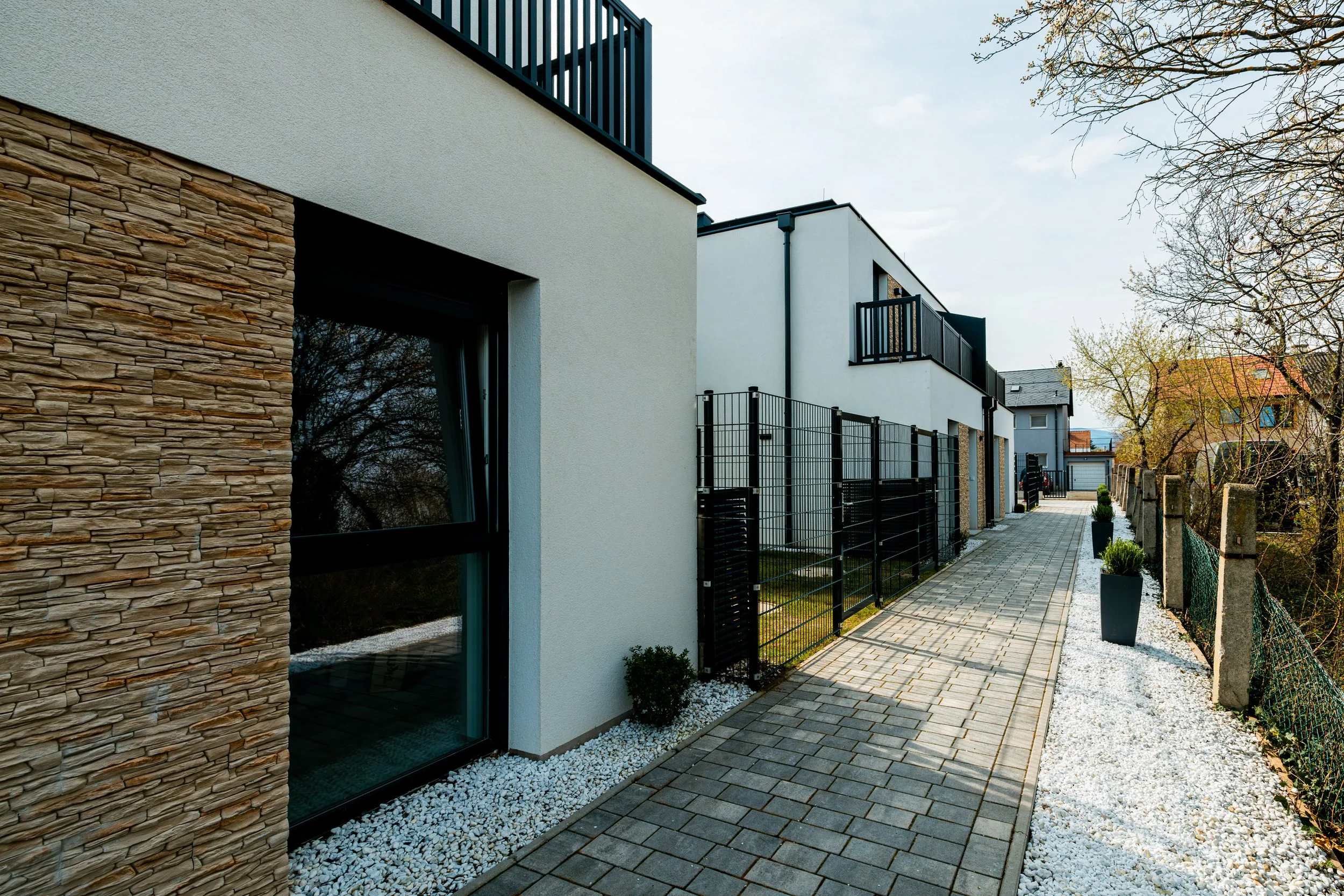 Moderne Mehrfamilienhäuser mit weiß gestrichenen Wänden, schwarzen Zäunen, einem Gehweg mit Pflastersteinen, kleinen Bäumen in Pflanzkübeln und einem bewölkten Himmel.