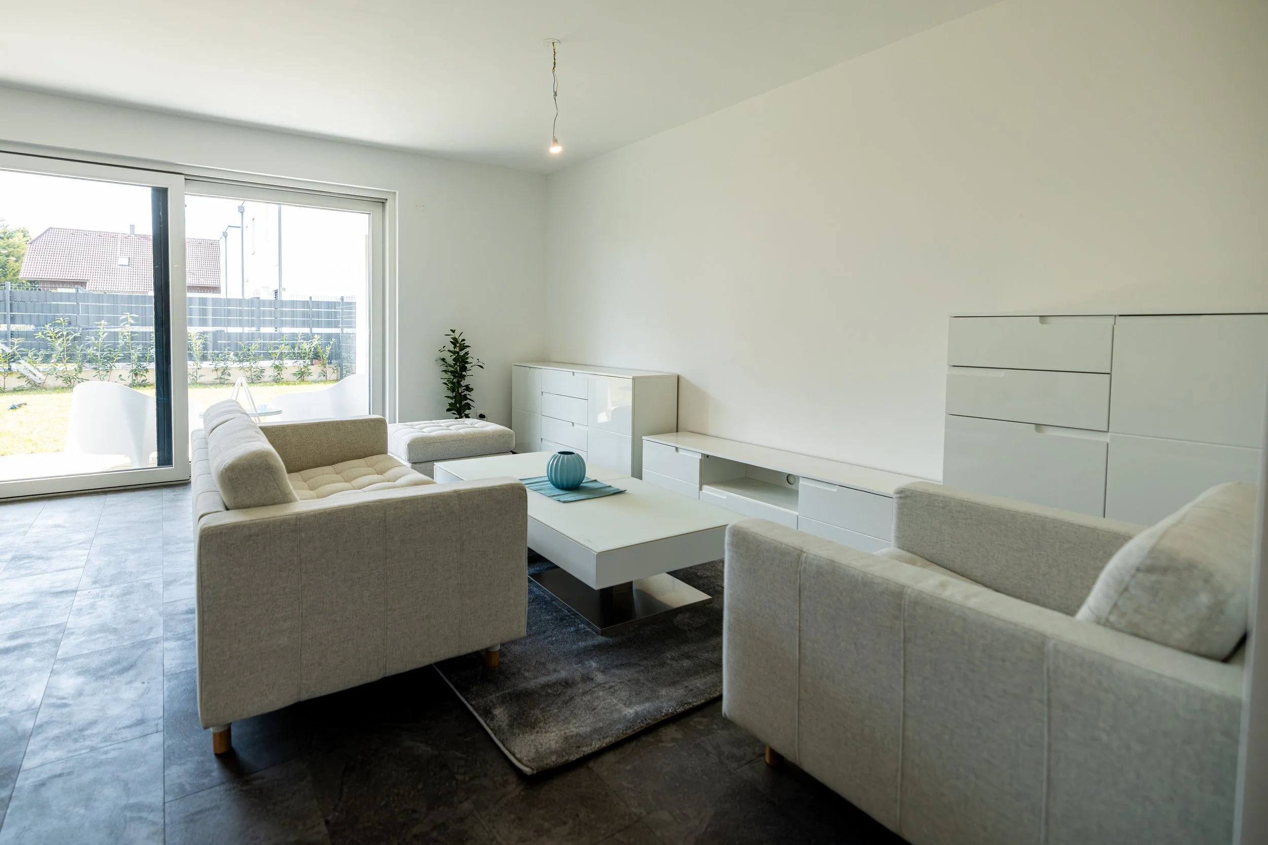 Helles Wohnzimmer mit weißen Sofas, Couchtisch, großen Fenstern, Blick auf Garten, weiße Möbel, minimalistische Einrichtung.