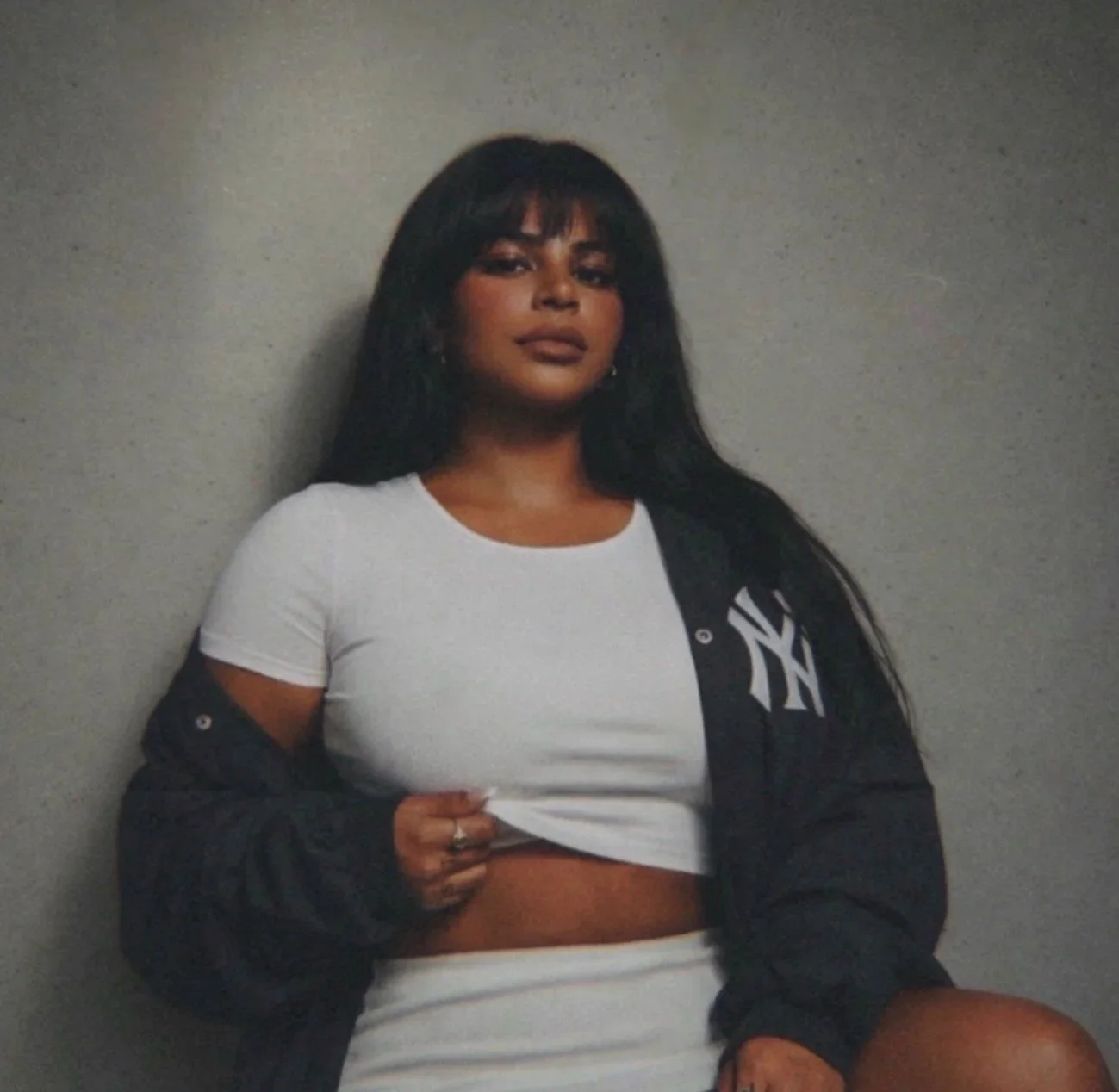 A woman with long dark hair and bangs, wearing a white crop top and a black jacket with a New York Yankees logo, posed against a plain gray wall.