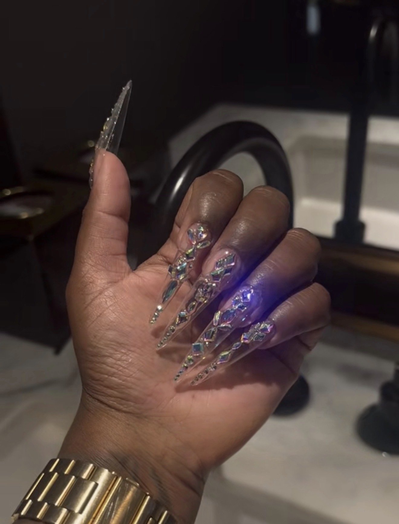 Hand with long, clear, jeweled nails and gold watch, holding a small glass object, with a piano in the background.