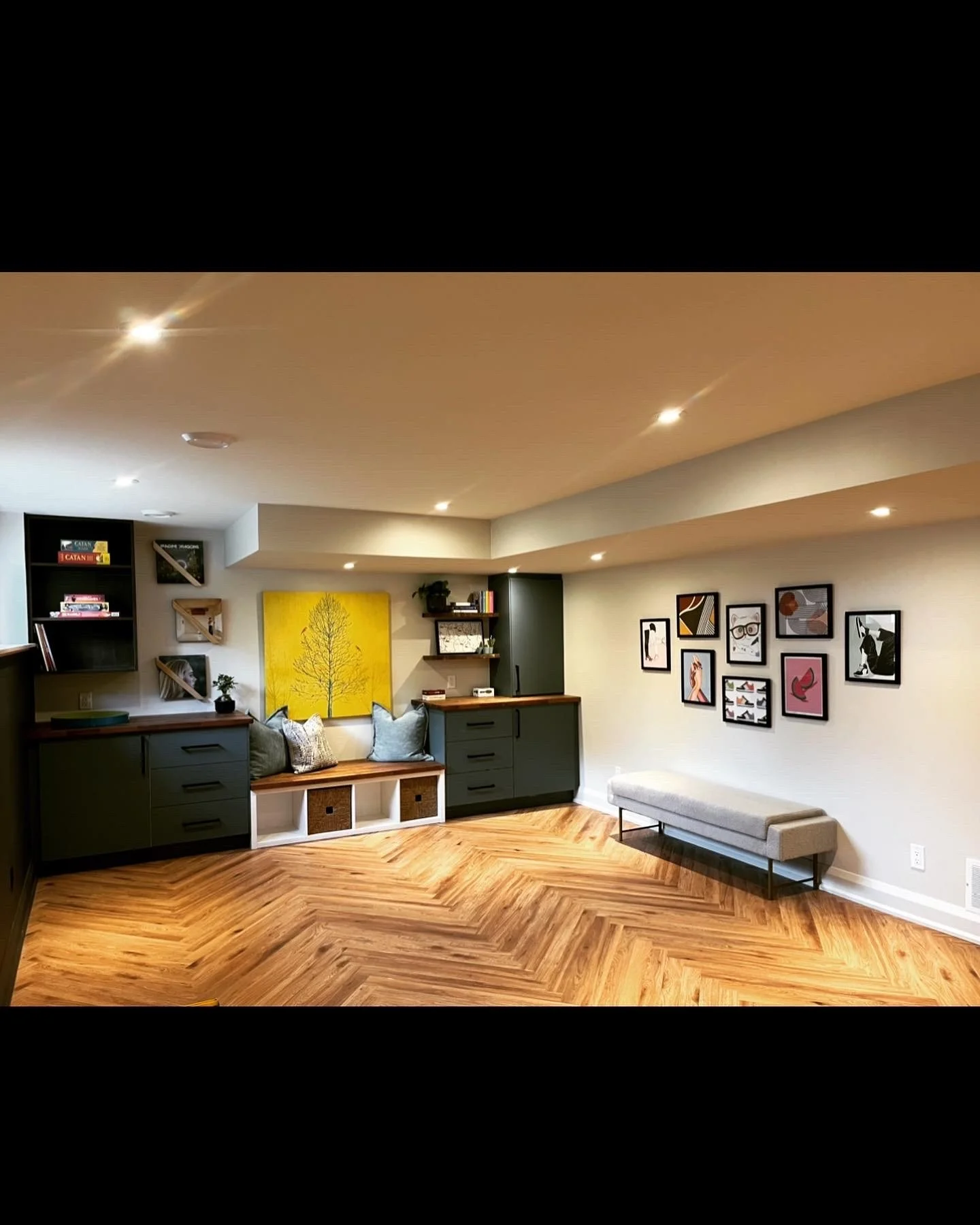 Interior view of a living room with wooden chevron-patterned flooring, built-in storage with dark green cabinets and shelves, a yellow artwork with a tree, a seating nook with cushions, a gray bench, and framed wall art, illuminated by recessed ceili