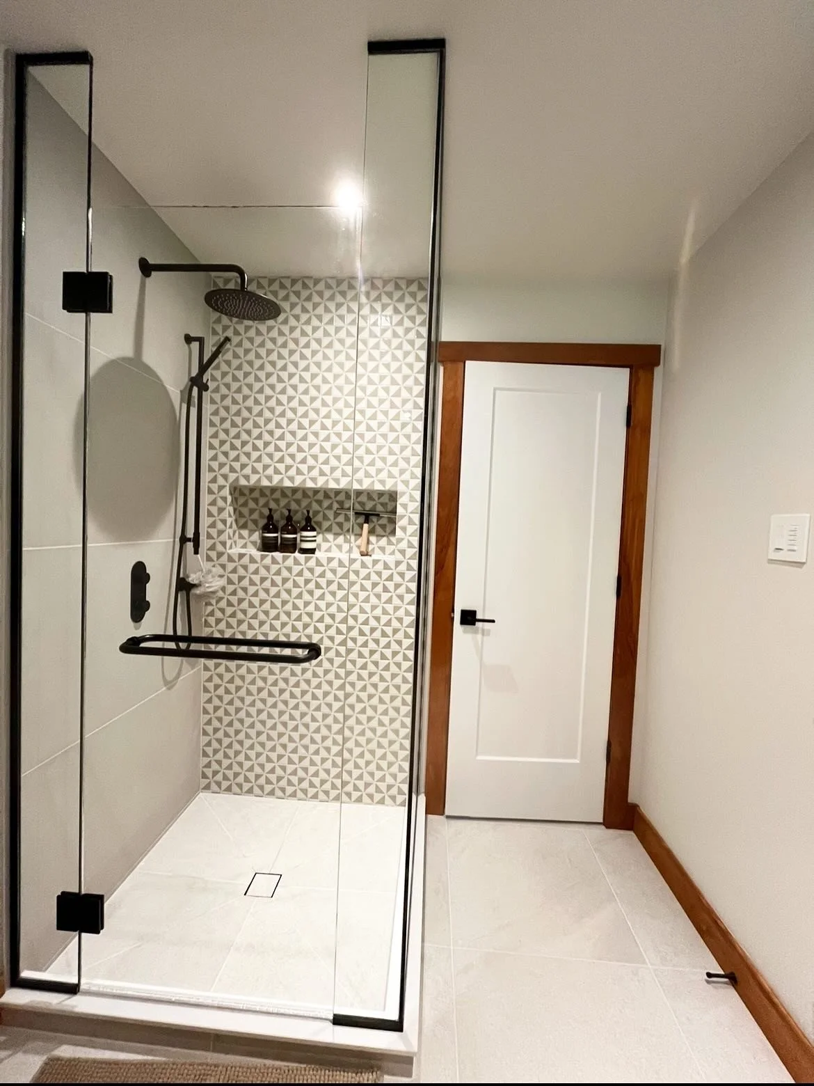 Bathroom with a walk-in shower enclosed by glass, featuring black fixtures, geometric wall tile, a built-in shelf with bottles, a wooden door, and a white wall with wood trim.