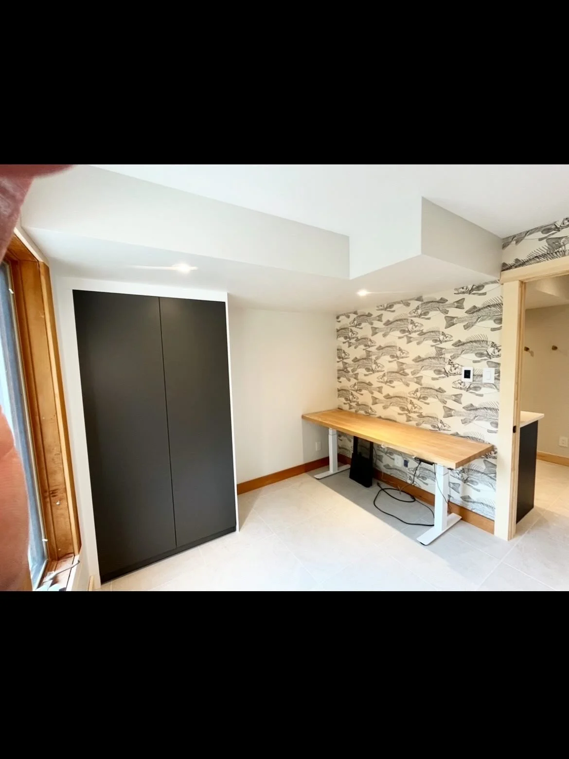 Part of a room with a black wardrobe, a wood desk with a black monitor underneath, and wallpaper with fish illustrations on the wall.