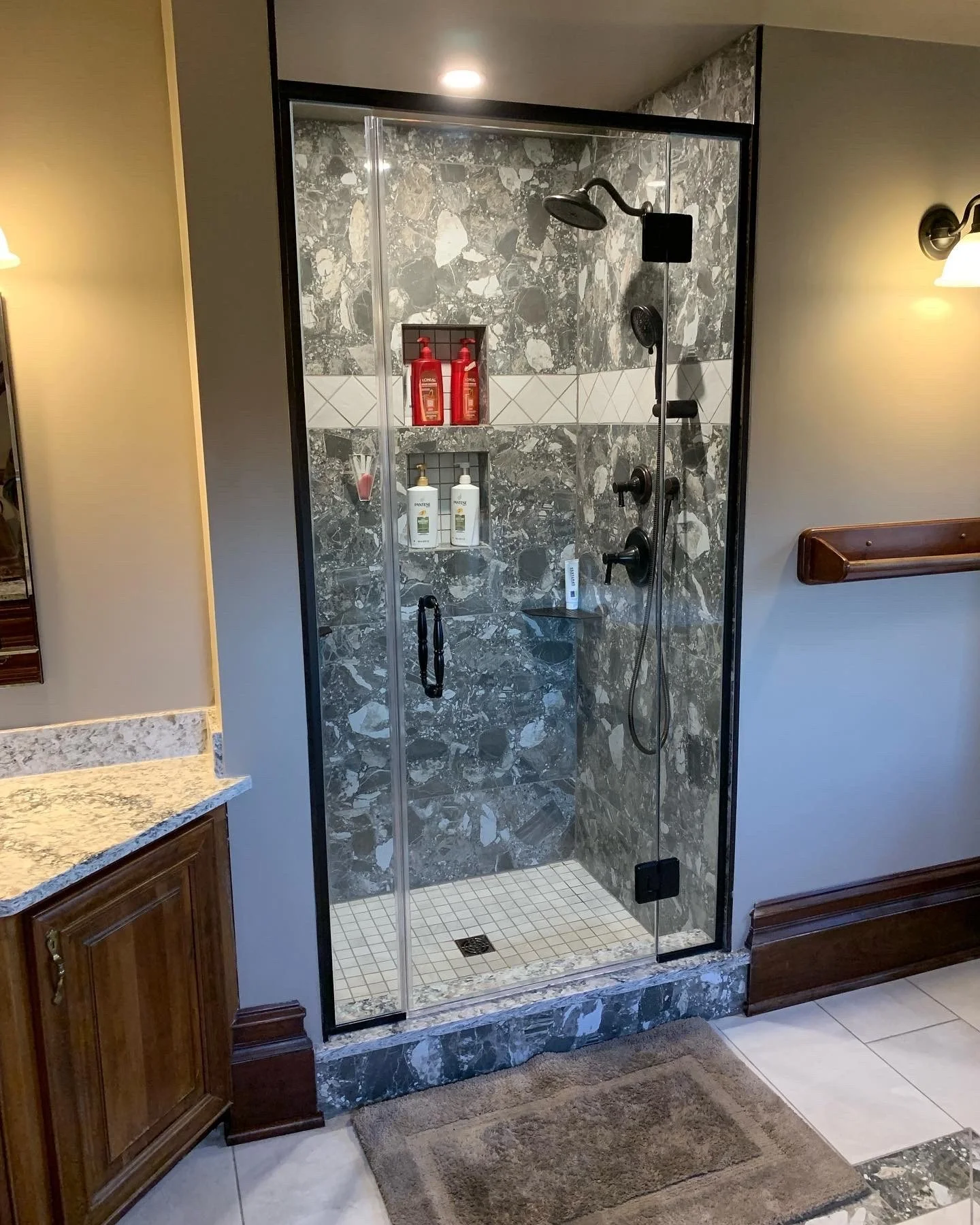 Inside a bathroom shower with gray marble walls and a glass door, containing red and white shampoo and soap bottles, showerhead, and a small shelf with toiletries, adjacent to a granite-topped sink and wooden cabinetry.