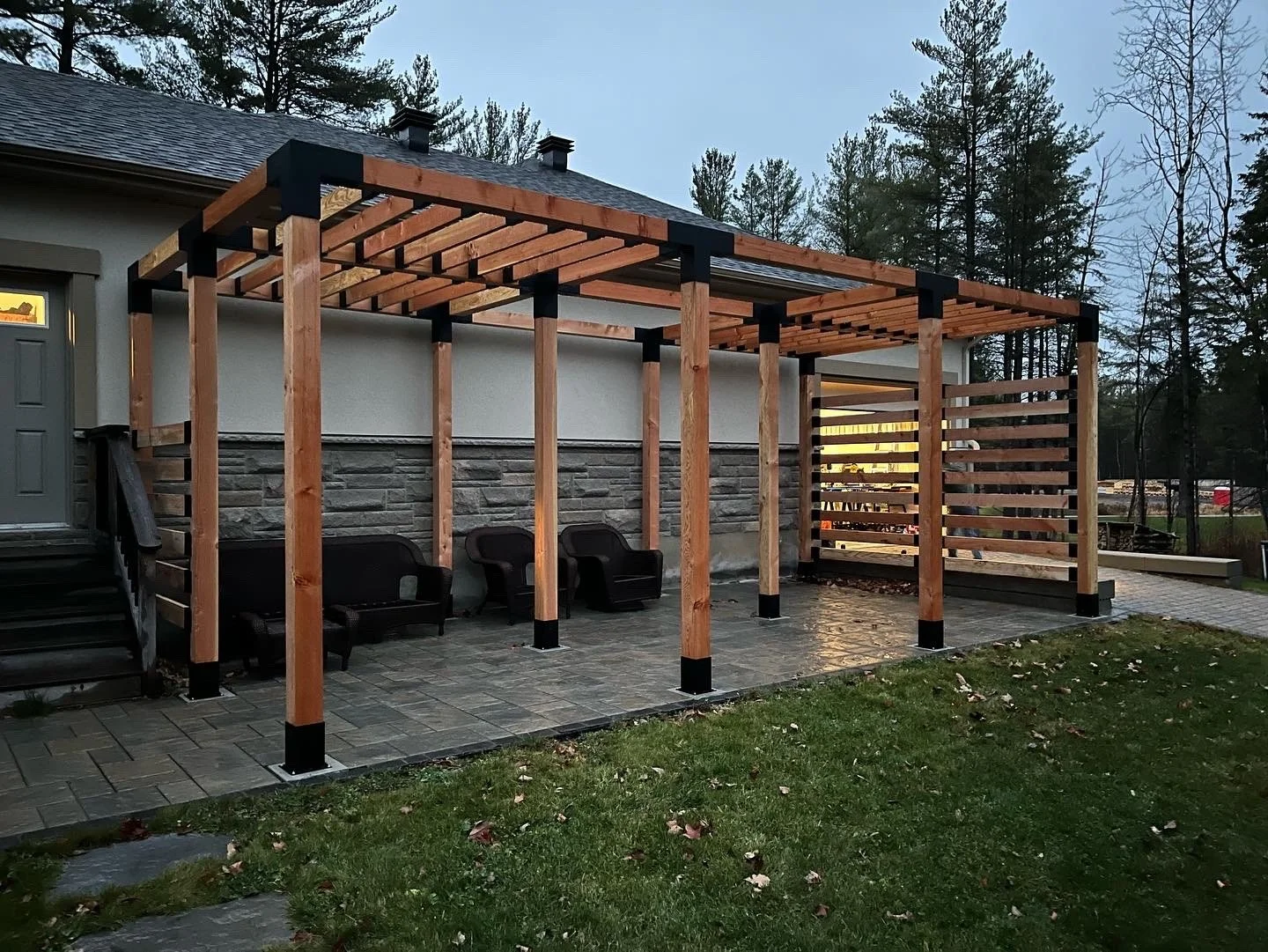 Wooden pergola under construction on a paved patio with black benches, next to a house with gray door and stone siding, in a backyard with grass and trees.