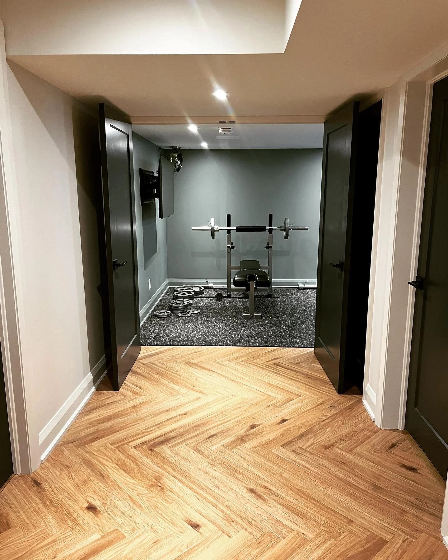 A home gym area with a weight bench, barbell, and weight plates on a rubber mat, visible through open black doors from a room with wooden flooring.