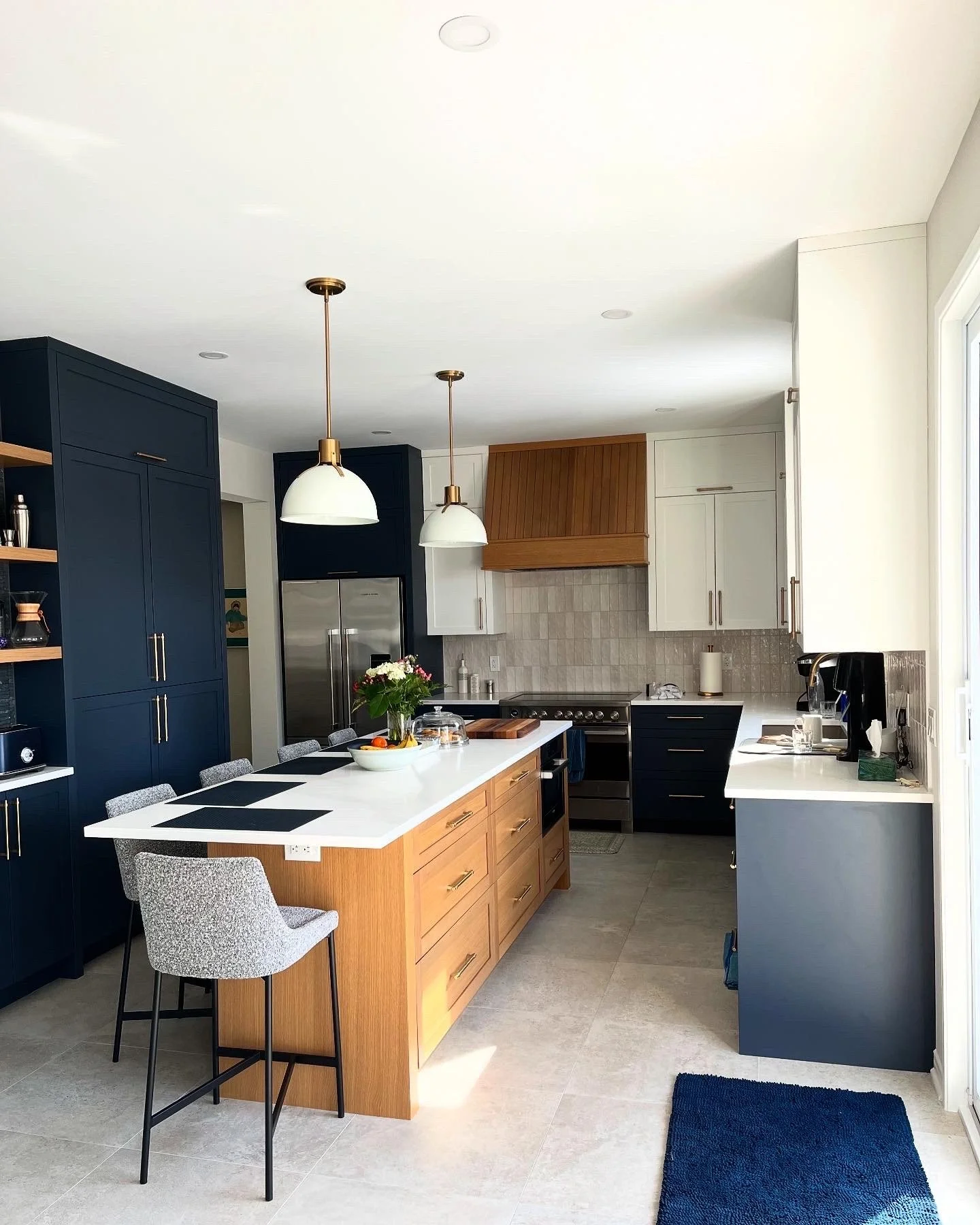Modern kitchen with navy blue, white, and wood cabinetry, a central island with a white countertop, bar stools, pendant lights, and a stainless steel refrigerator.