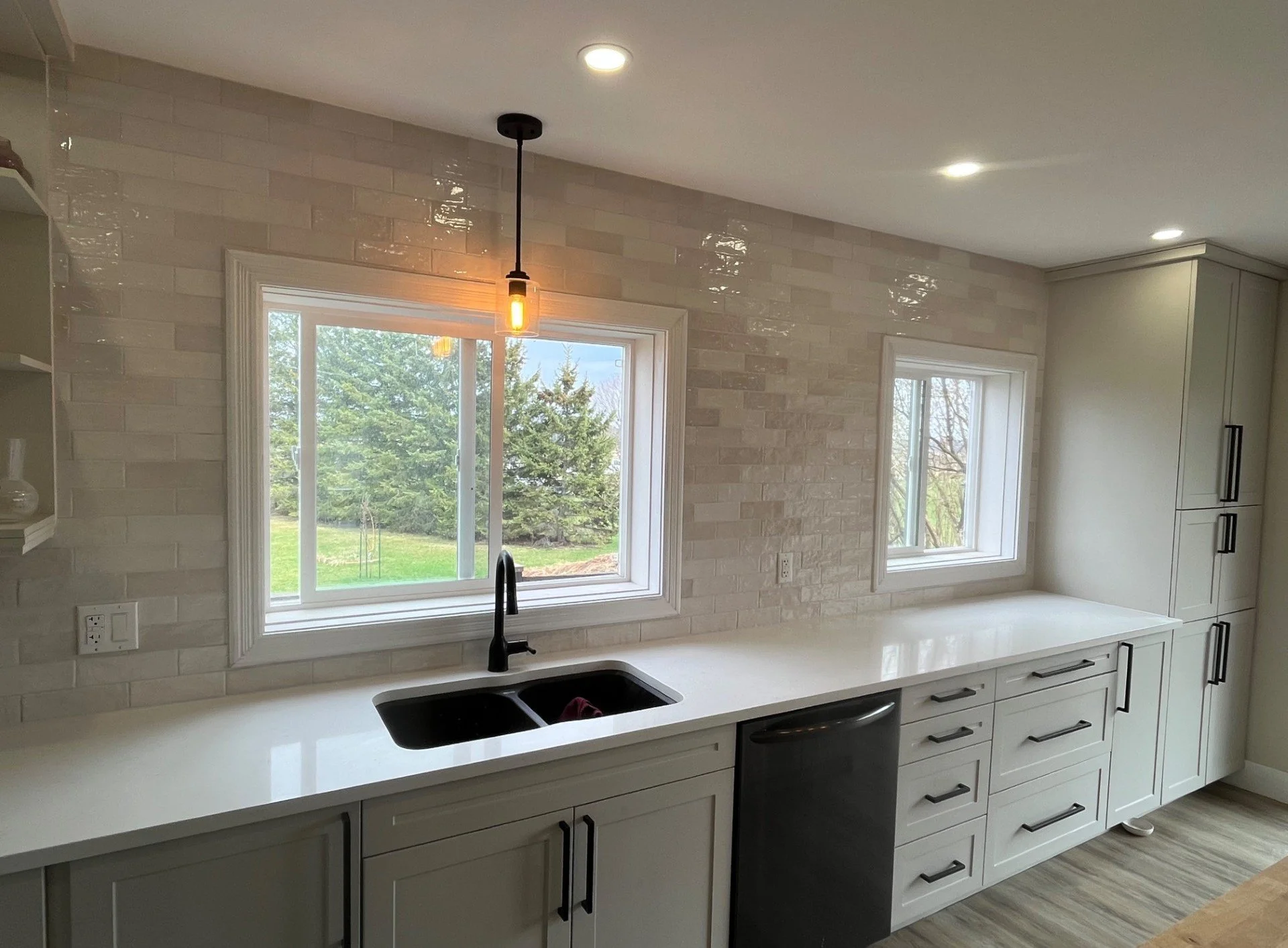 Modern kitchen with white cabinets, black handles, a black sink, and a white countertop   windows overlooking a green outdoor area, minimalistic decor, natural light, with two small windows, wood-look flooring, and a hanging light bulb fixture.