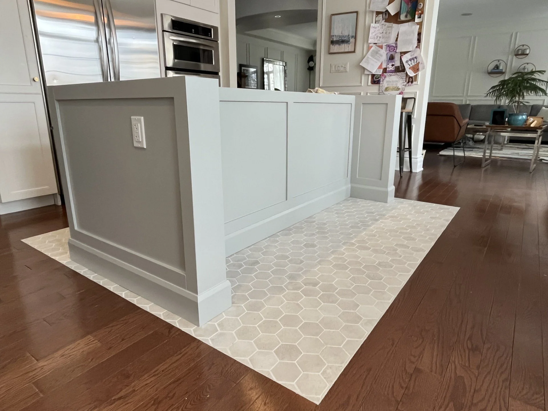 Kitchen island with light gray cabinetry on a patterned beige and white hexagon tile floor, in a modern open-plan living space with hardwood floors, a wall with drawings and notes, and a living room with couches and plants in the background.