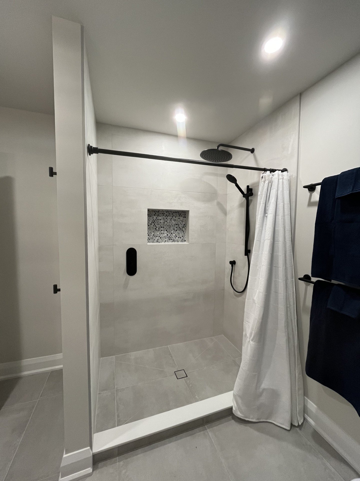 Modern walk-in shower with a rainfall showerhead, handheld shower, white tile walls, a built-in niche, and a white shower curtain, with navy towels hanging on the side.