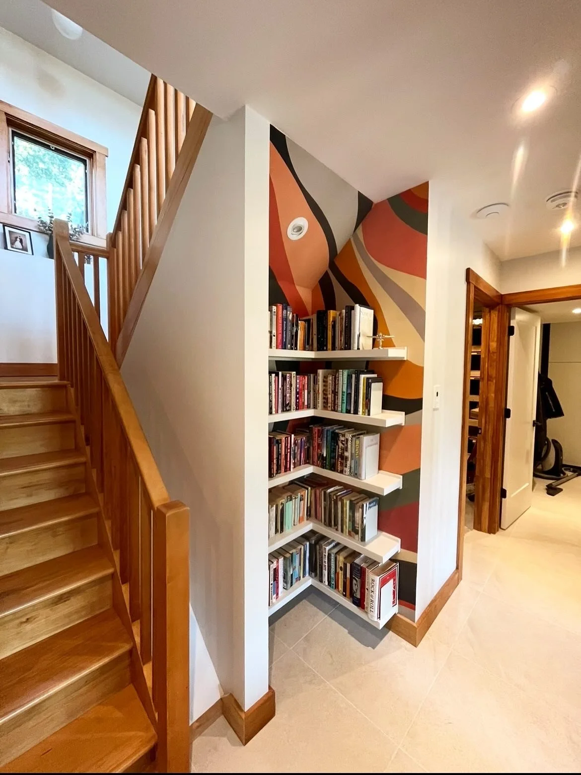 A corner of a room with wooden stairs on the left, a colorful abstract mural on the wall, and a white bookshelf filled with books below the mural. A small window is visible at the top of the stairs.