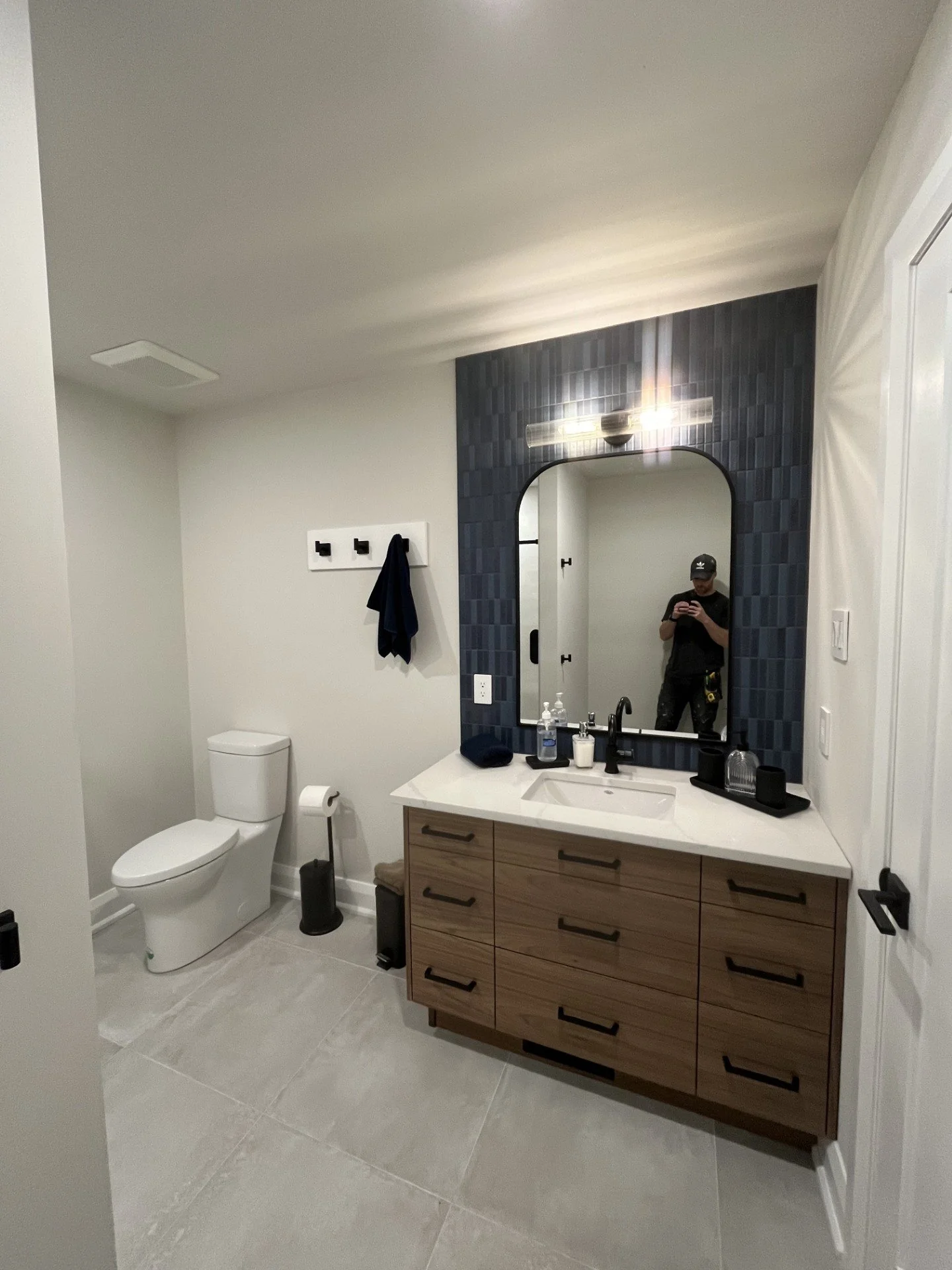 Modern bathroom with a wooden vanity, white countertop, large mirror, and navy blue textured tile wall behind the mirror. Contains a toilet and black towel hooks with a towel hanging. Reflects a person taking a photo with a smartphone.
