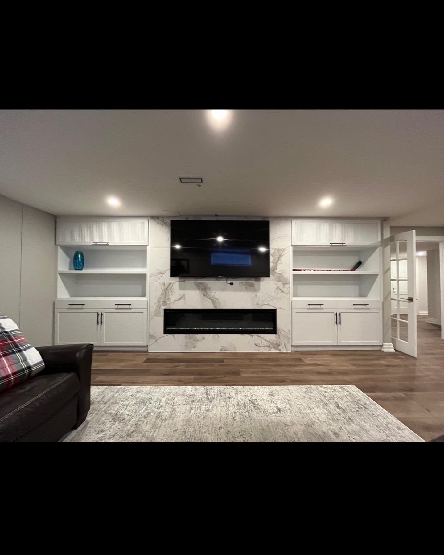 Living room with white built-in shelves, a mounted TV, marble accent wall, fireplace, hardwood flooring, and a gray area rug. Part of a black leather sofa with a plaid pillow is visible.