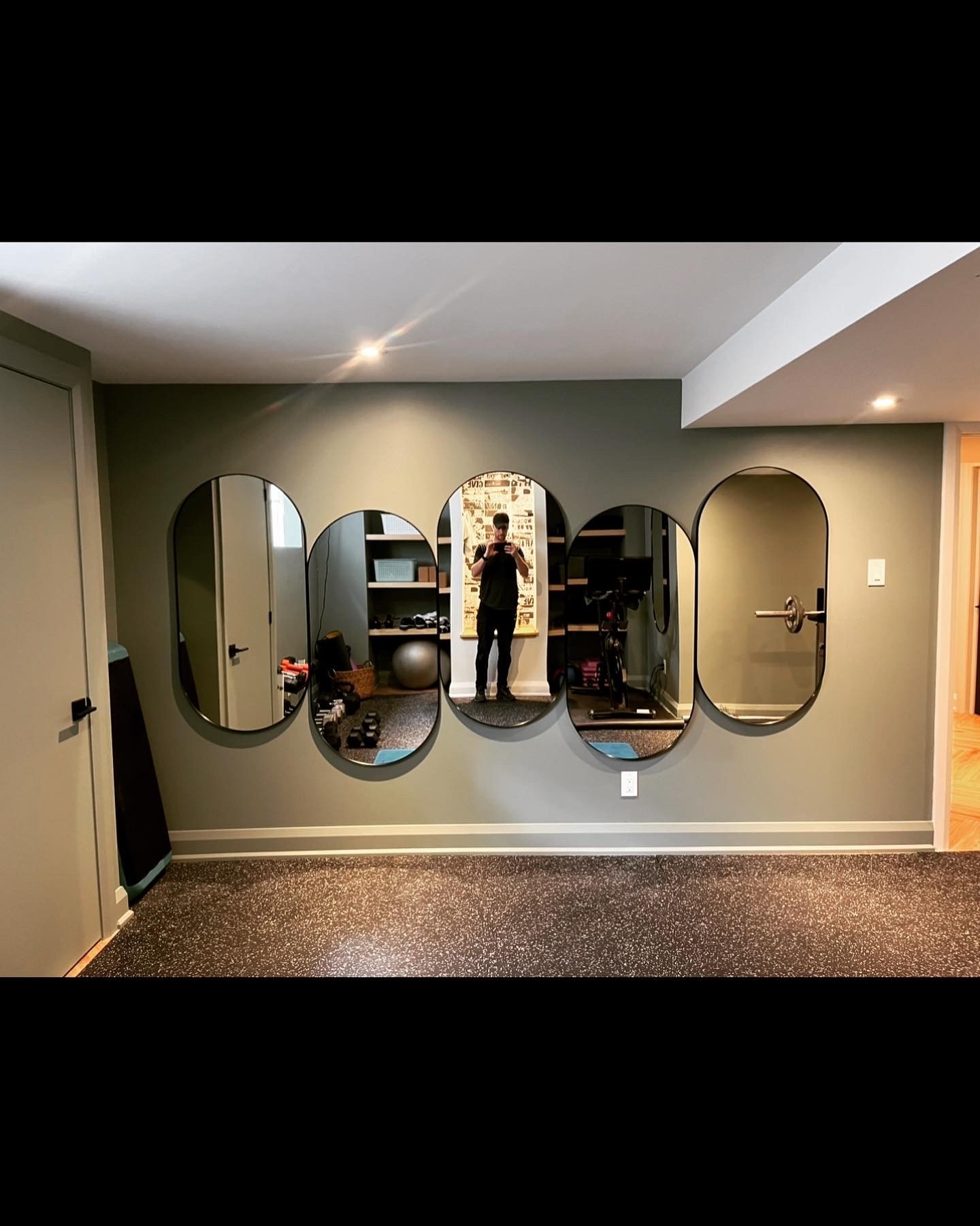 A wall with a series of five oval mirrors, a person taking a photo, and gym equipment in the reflection.