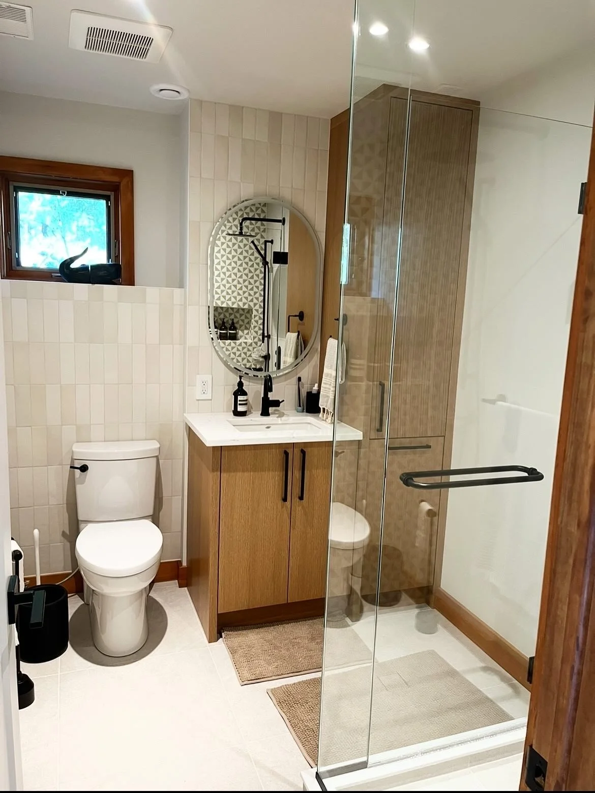A modern bathroom with a toilet, a wooden vanity with a sink, a mirror, and a glass-walled shower stall. There is a small window and a black sculpture on the window sill.