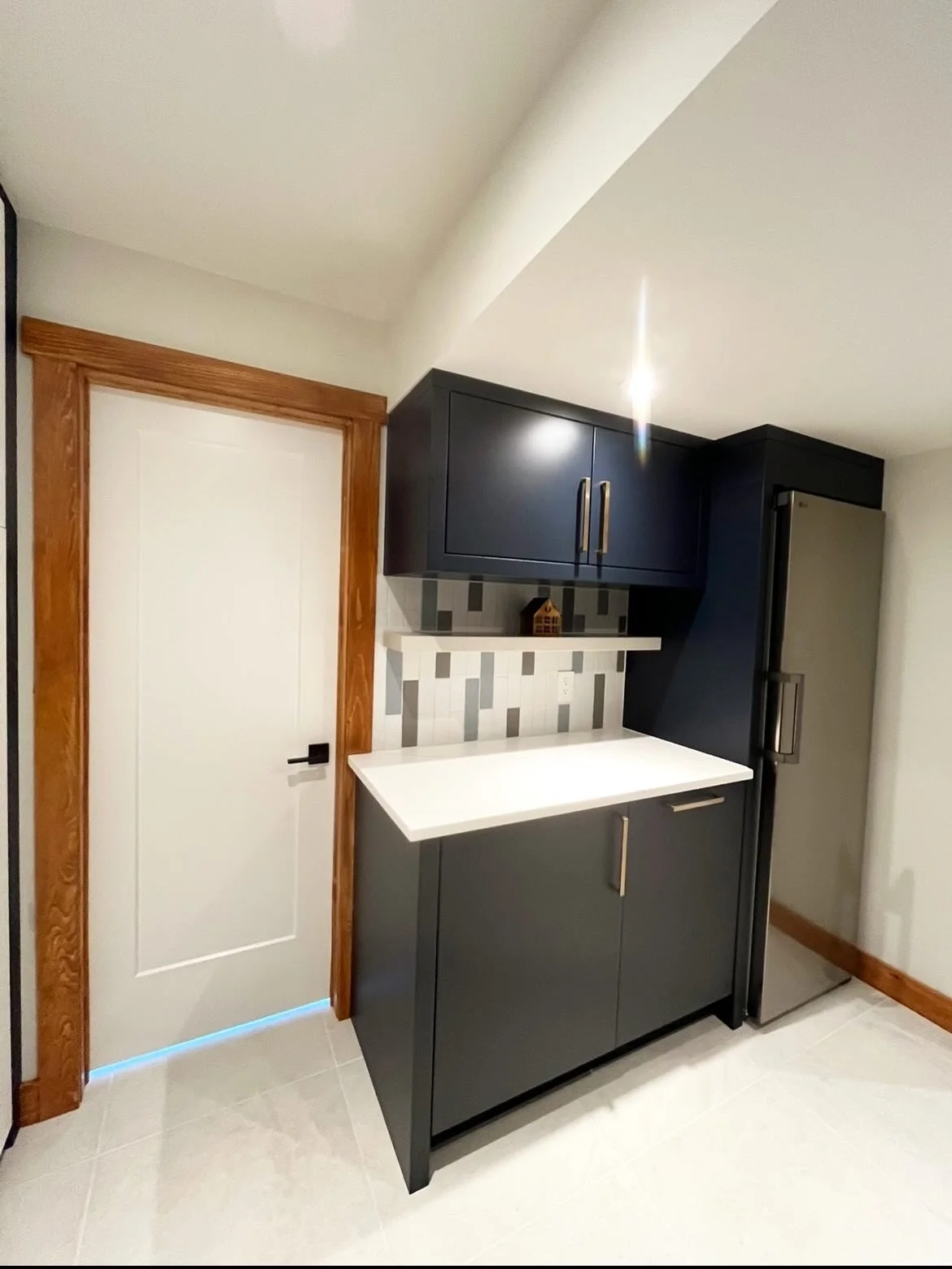 Contemporary kitchen with black cabinetry, a white countertop, gray and white backsplash tiles, a small shelf, and a stainless steel refrigerator.