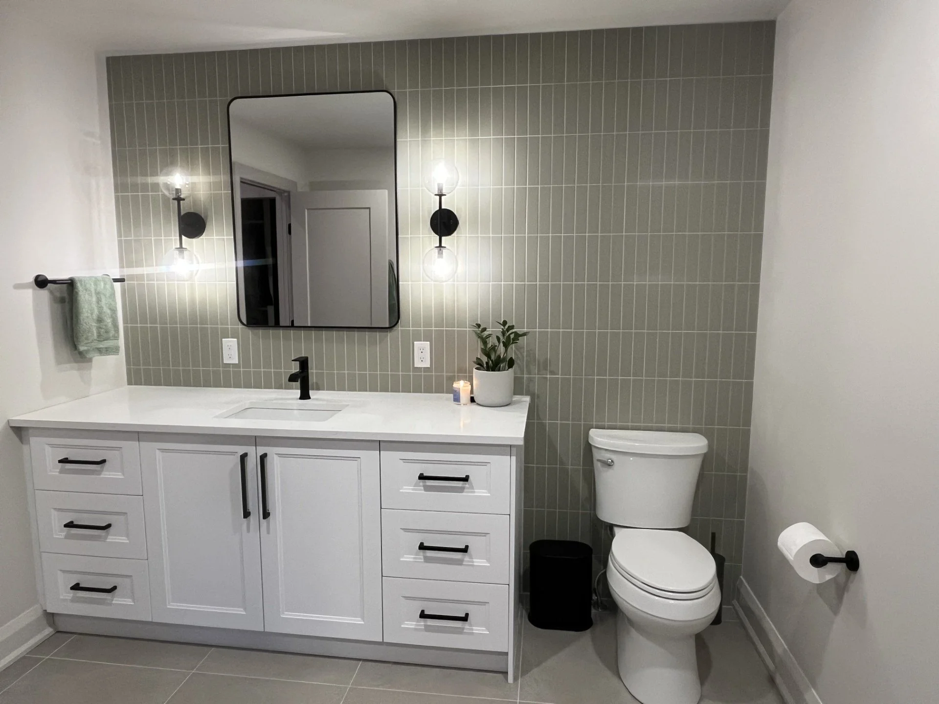 Modern bathroom with white vanity, black faucet, rectangular mirror, wall-mounted lights, potted plant, candle, toilet, black waste bin, and beige tiled wall with vertical tiles.