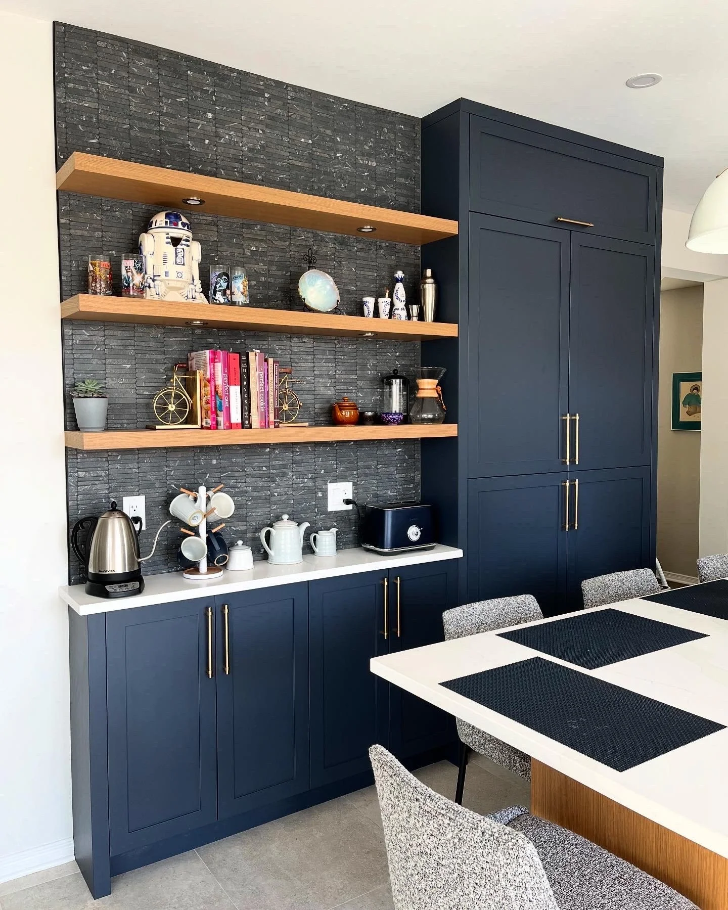 Kitchen cabinet with open shelves holding decorative items, books, and kitchenware, against a black textured wall with a dining table nearby.