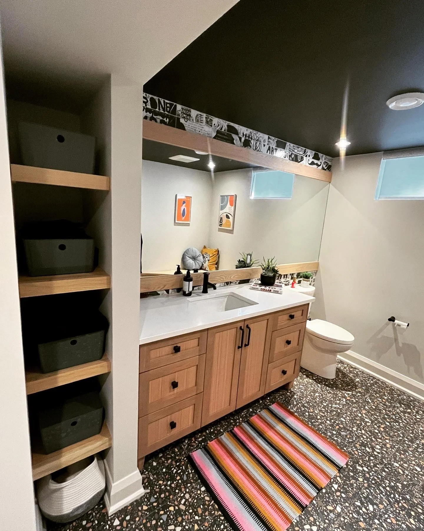 A bathroom with a wooden vanity, a mirror, and a toilet. The floor is speckled, and there are colorful art pieces on the wall. A striped rug is on the floor, and there are plants and toiletries on the vanity.