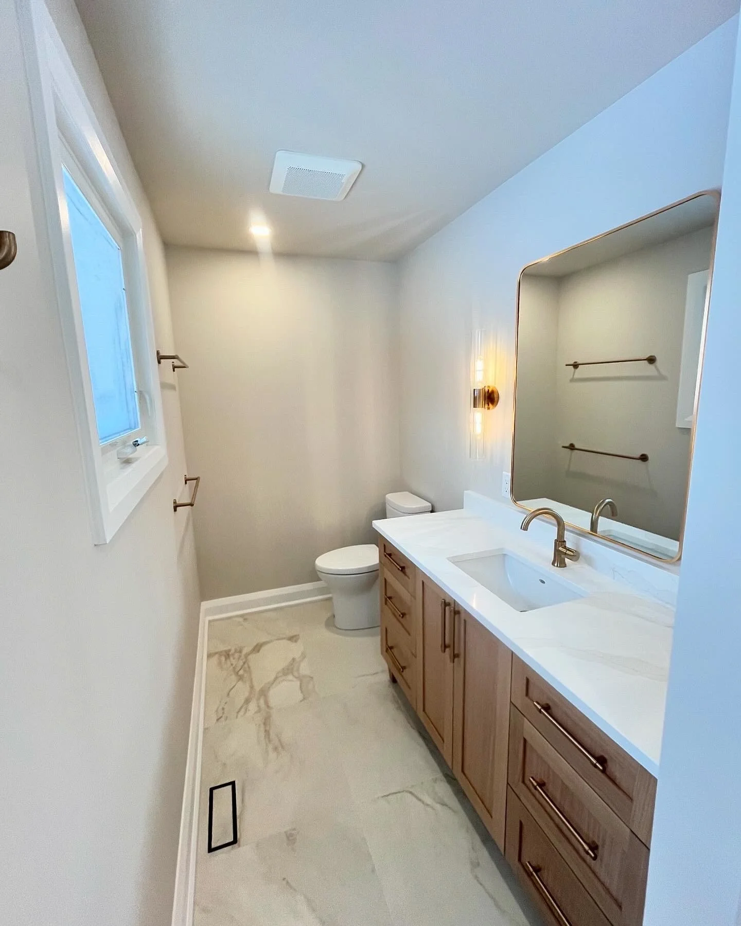 Modern bathroom with a wooden vanity, white countertop, large mirror, single sink, wall-mounted light, toilet, window, and marble floor tiles.