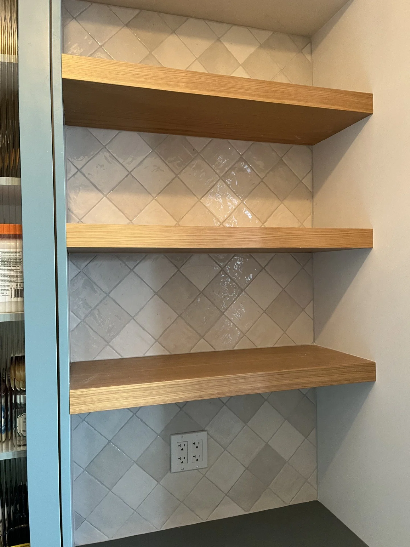 Empty wooden shelves in a kitchen next to a wall with beige diamond-shaped tiles and a power outlet.