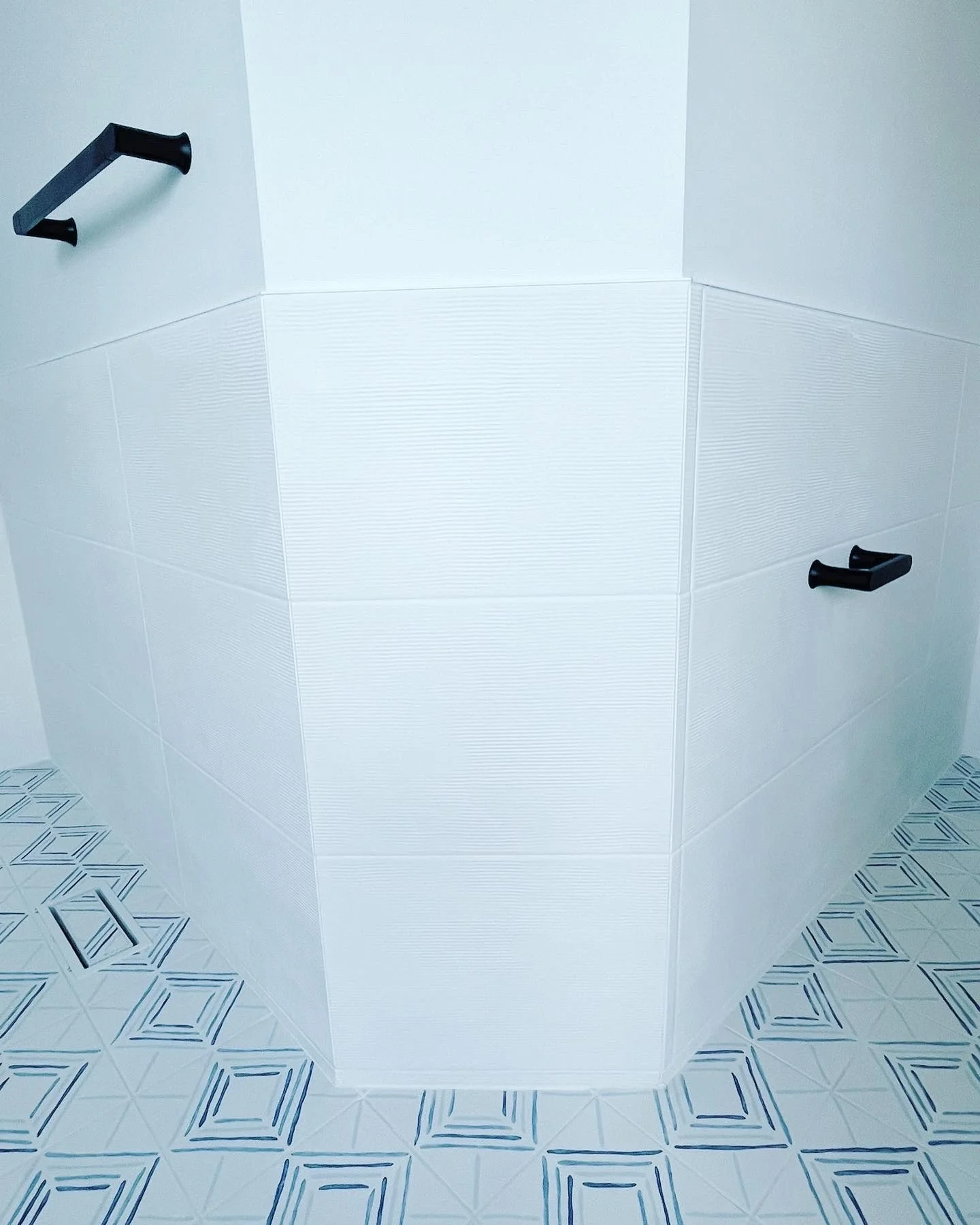 Corner of a white tiled bathroom with black grab bars and patterned floor tiles.