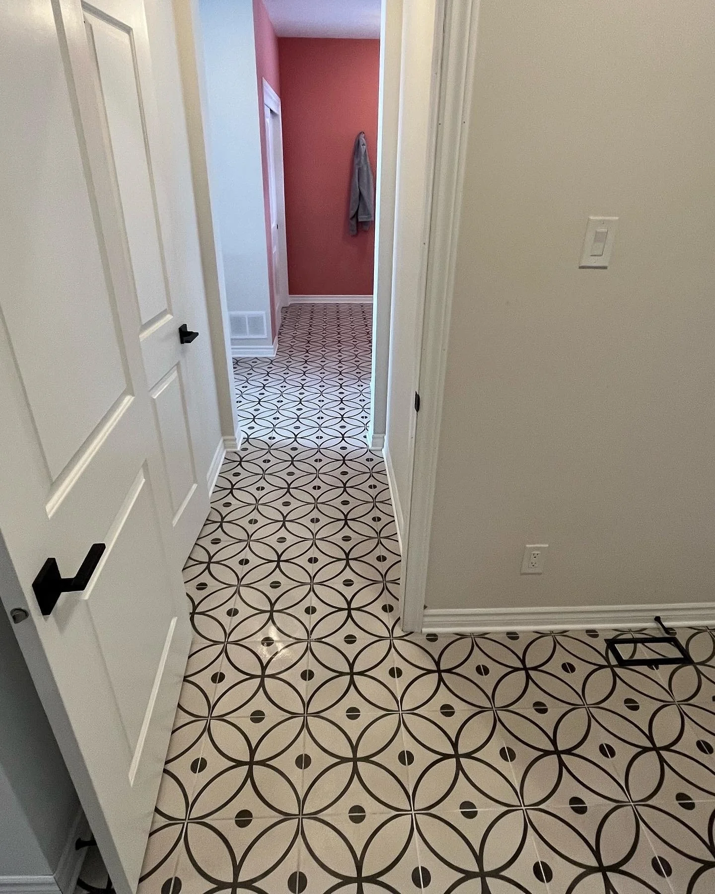 View of an indoor hallway with patterned tile flooring, white doors, and a pink accent wall at the end. A gray garment hangs on a hook on the pink wall.