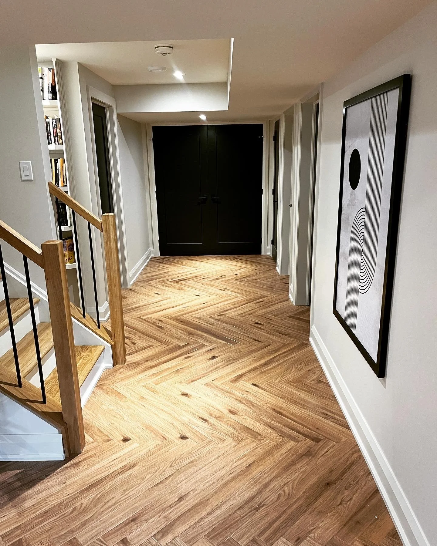 Interior view of a hallway with wooden herringbone-pattern floor, a staircase with wooden handrail, white walls decorated with framed artwork, built-in bookshelf, and black double doors at the end.