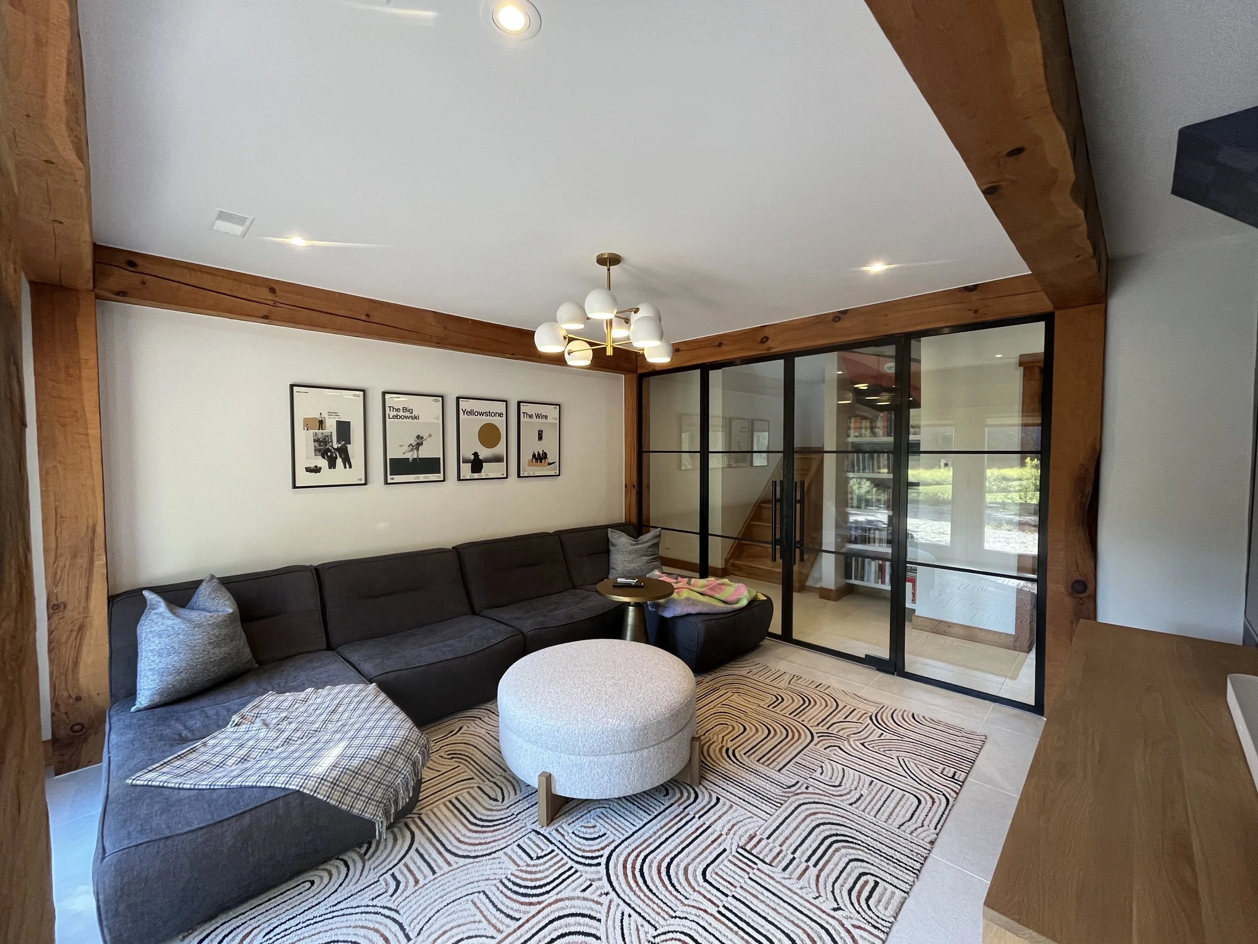 Living room with a black sectional sofa, modern artwork on white wall, wooden beams and glass sliding door, beige rug with circular patterns, ceiling light fixture, and wooden staircase in background.