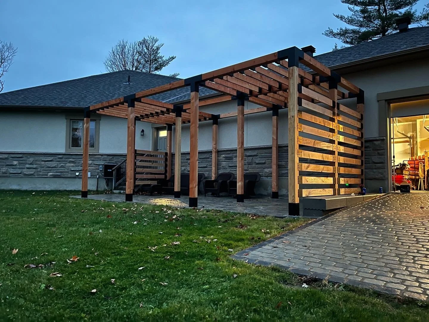 A partially constructed wooden pergola outside a house with a stone and stucco exterior. The structure has vertical posts and horizontal beams, with some sections enclosed by horizontal slats. There are three black chairs underneath, and a cobbleston
