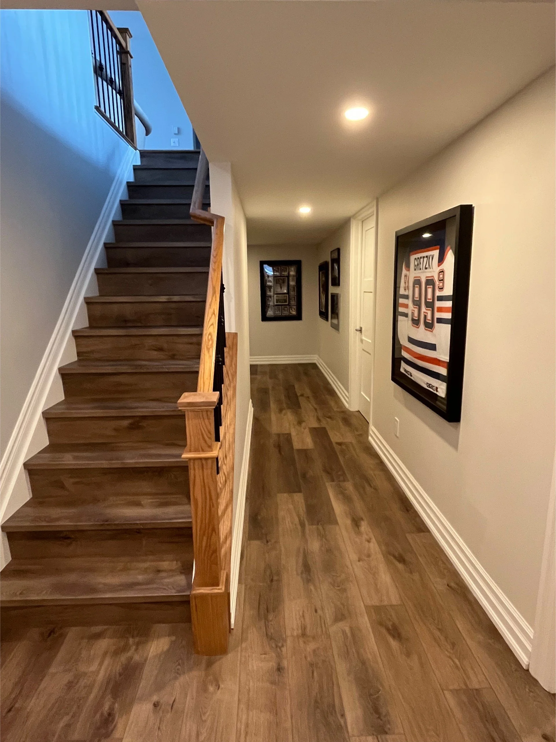 Interior view of a hallway with wooden flooring, framed sports jerseys on the wall, and a staircase with wooden steps and railing.