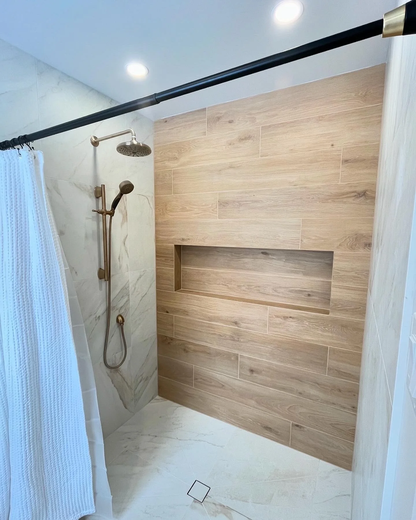 Modern walk-in shower with wood and marble tile walls, a built-in shelf, a brass showerhead, and a black curtain rod in a bathroom.