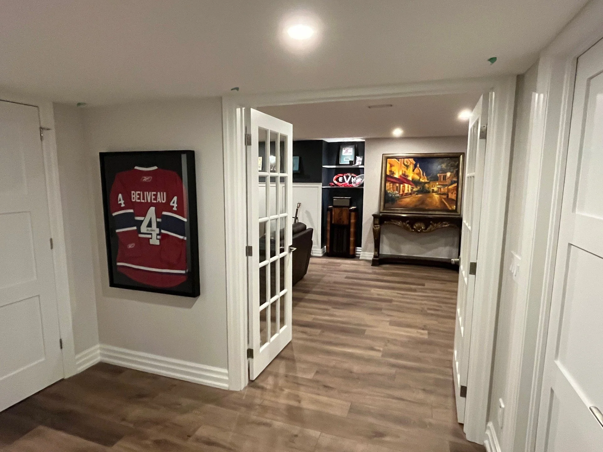 Interior view of a basement with hardwood flooring, white walls, and various decorations including a framed Montreal Canadiens hockey jersey, a framed painting, and a neon sign, with a doorway leading to a seating area.