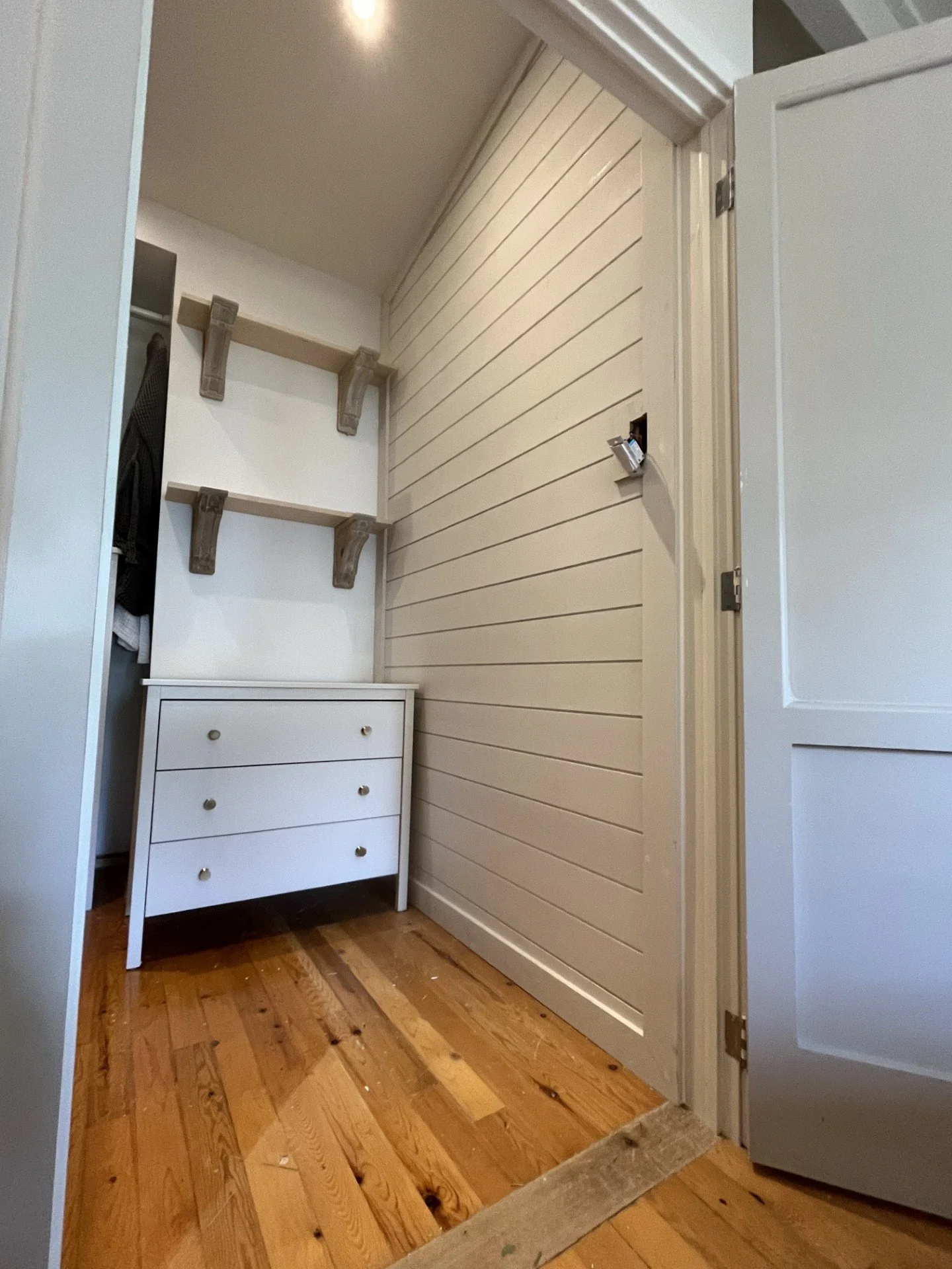 Small closet with white walls, a white chest of drawers, empty wooden shelves on wall, and its door slightly open, revealing wood flooring.