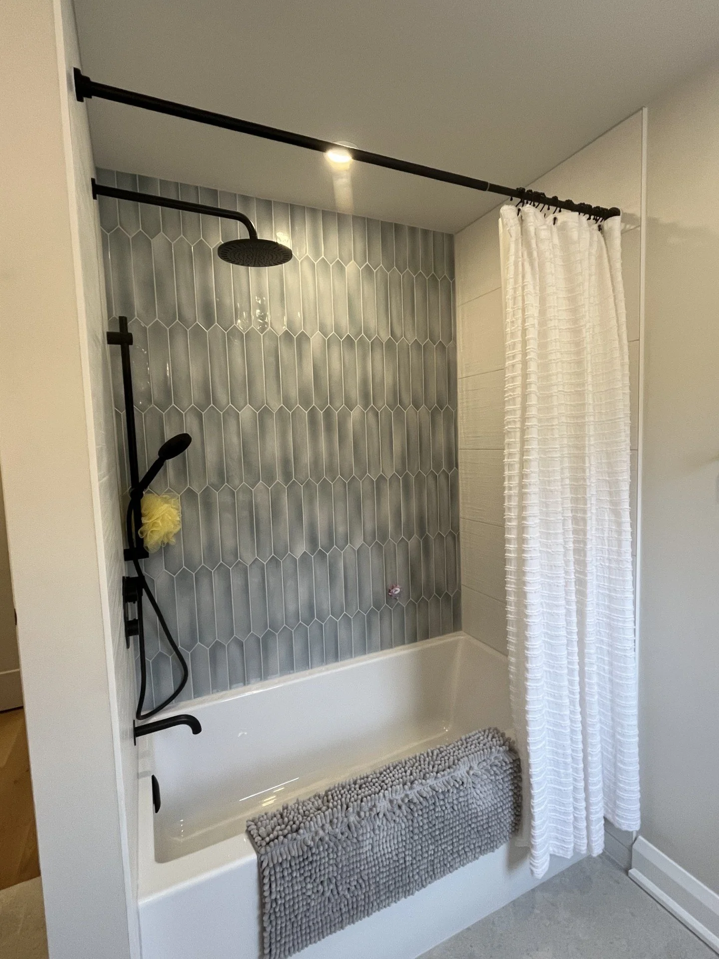 Modern bathroom shower with gray tile accent wall, black shower fixtures, white bathtub, white textured shower curtain, gray bath rug, and white walls.