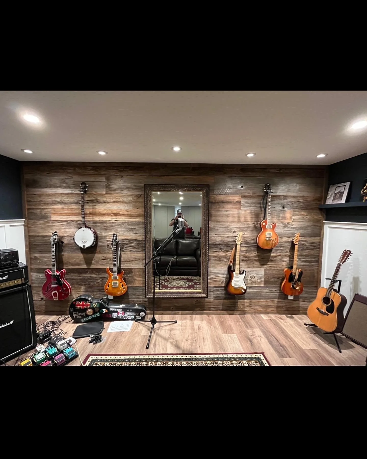 Music studio with six guitars hanging on a wooden wall, a mirror, and music equipment on the floor.