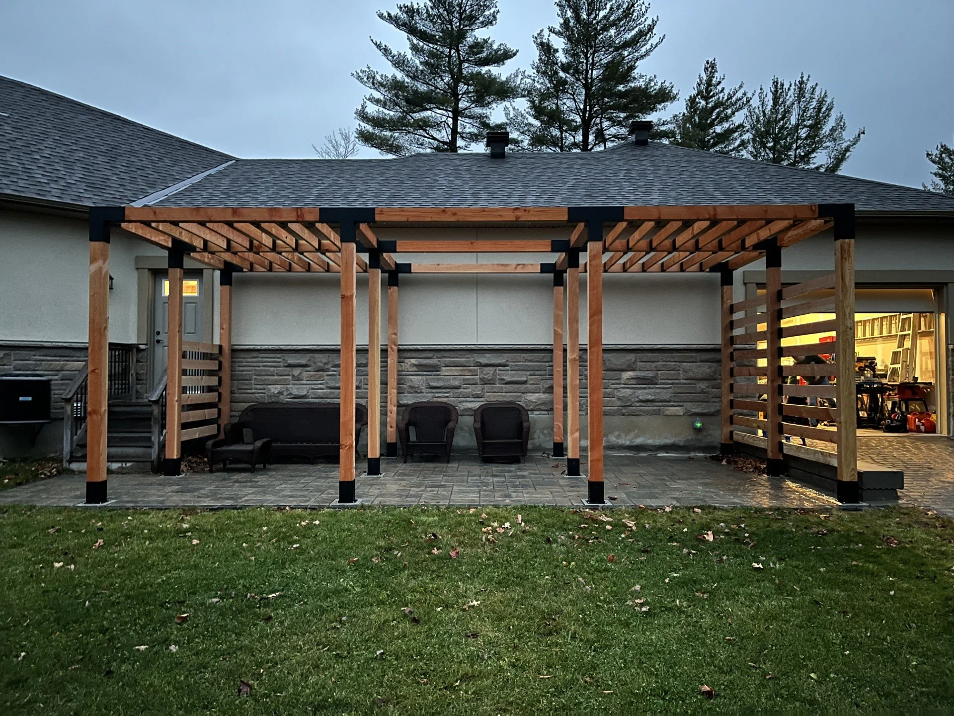 A backyard patio is under construction with a wooden pergola frame, partially completed. The patio floor is paved, and there are three black chairs and a black sofa placed along the house wall. The house has a sliding door with a room visible inside.