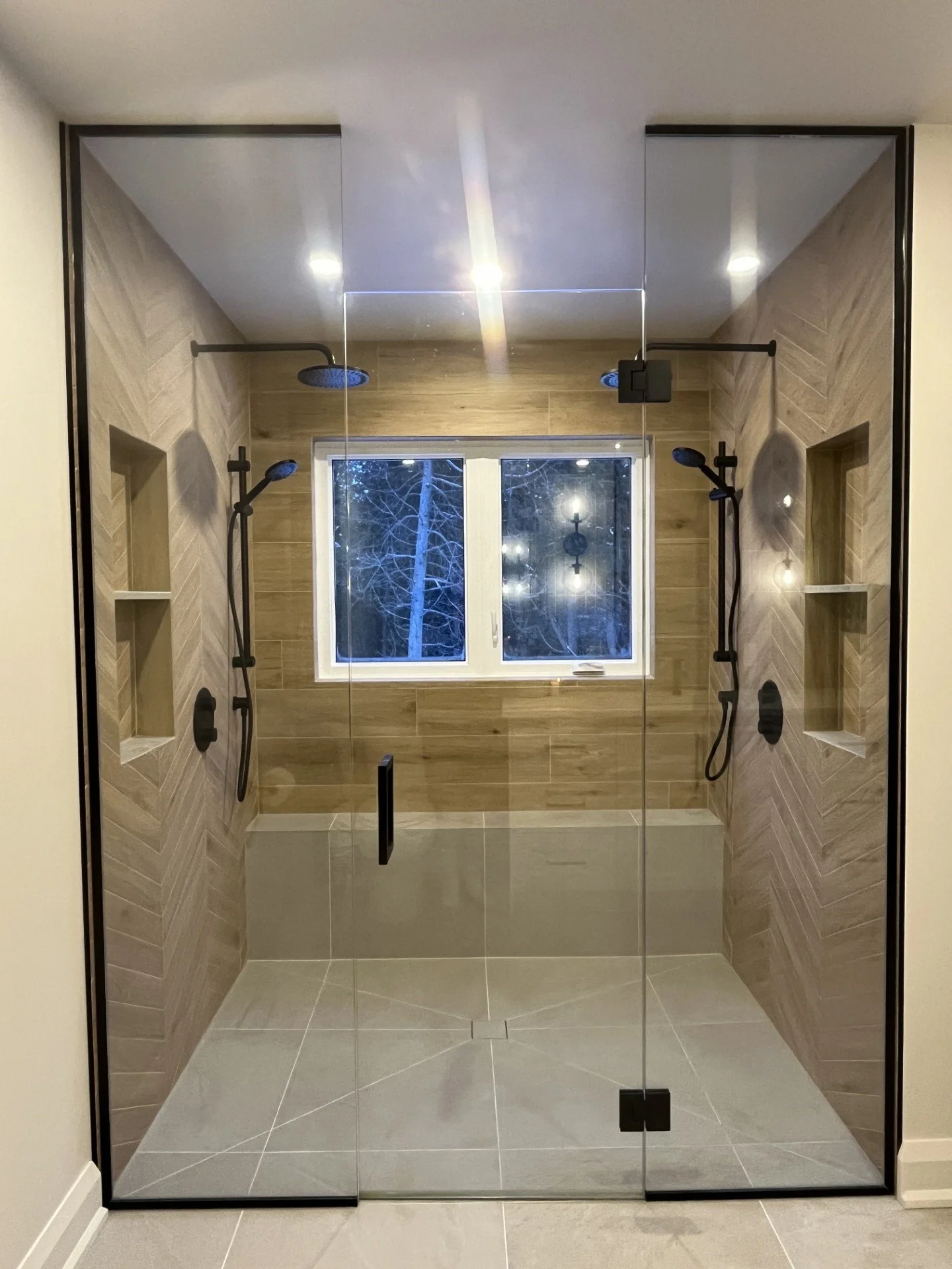 Modern walk-in shower with glass doors, two showerheads, built-in niches, a window, and wood-look wall tiles.