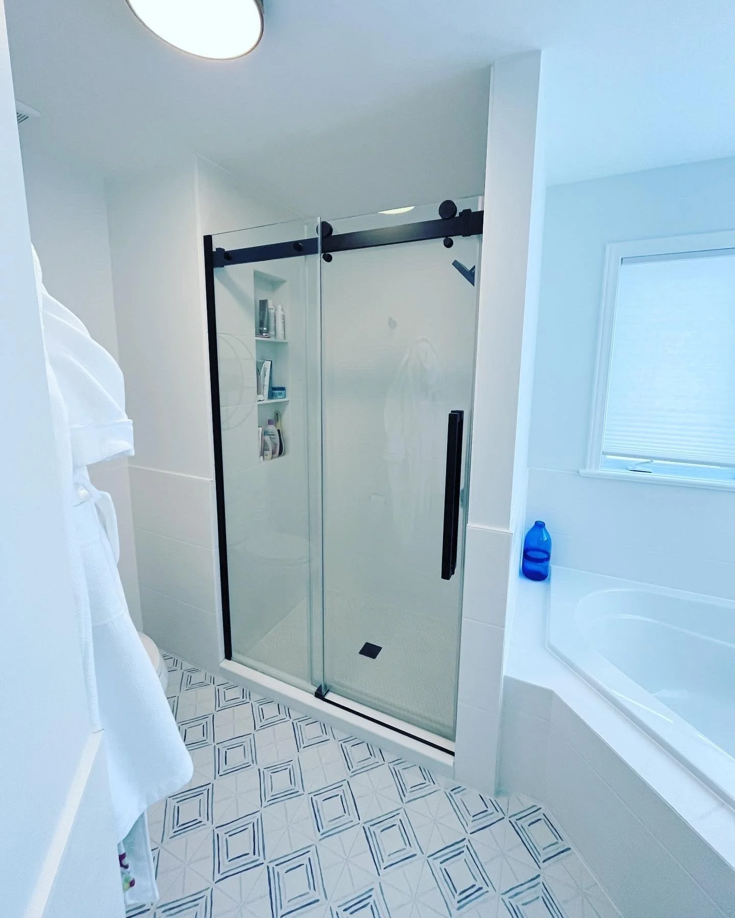 Bathroom with a glass shower door, a bathtub with a blue bottle nearby, a window with blinds, and white towels hanging.