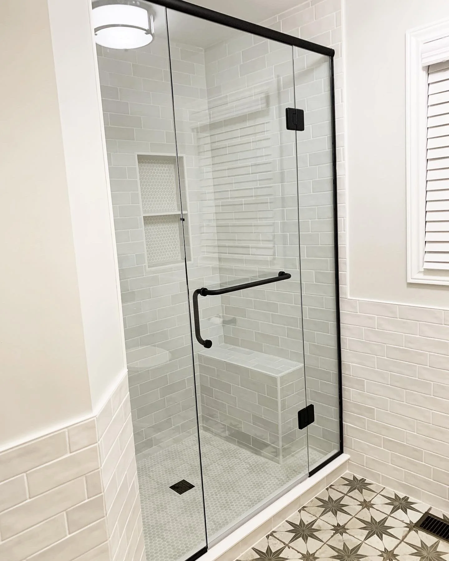 A modern glass shower enclosure with black hardware, white subway tile walls, a built-in niche shelf, and geometric patterned floor tiles.