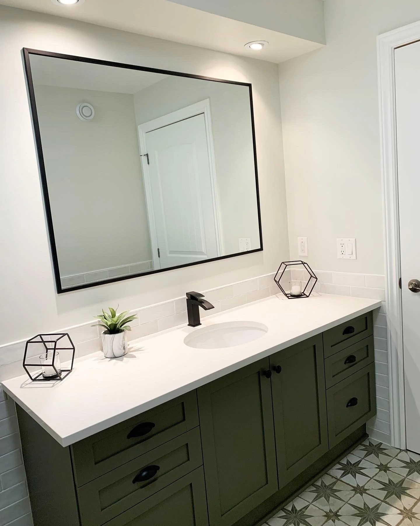 Bathroom with a large rectangular mirror, white countertop with a built-in sink, black faucet, green cabinet, white subway tile backsplash, decorative candles in geometric candle holders, and a small potted plant. Tiled floor with a geometric pattern