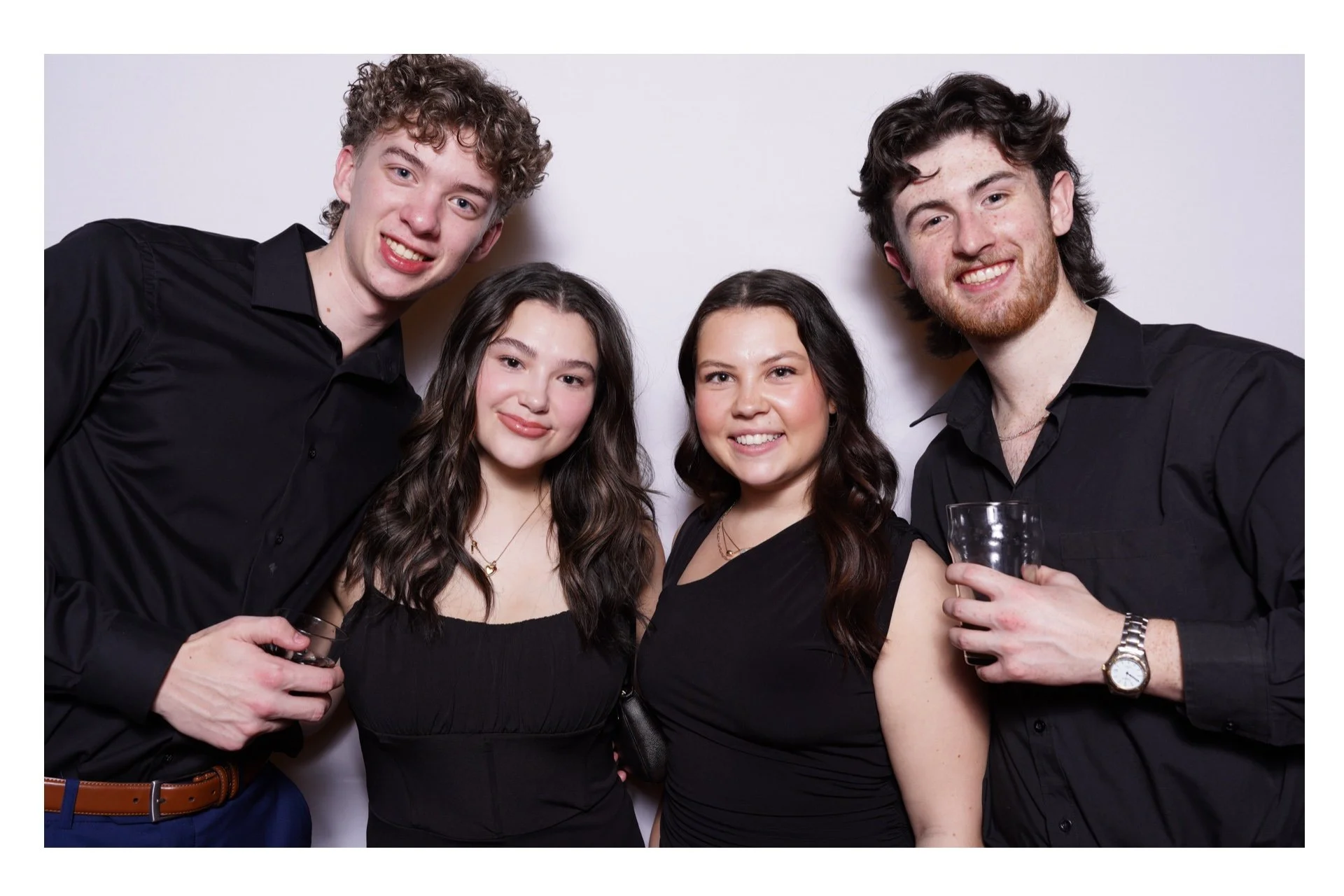 Group of four young adults at a party, smiling and holding drinks in front of a plain white background.