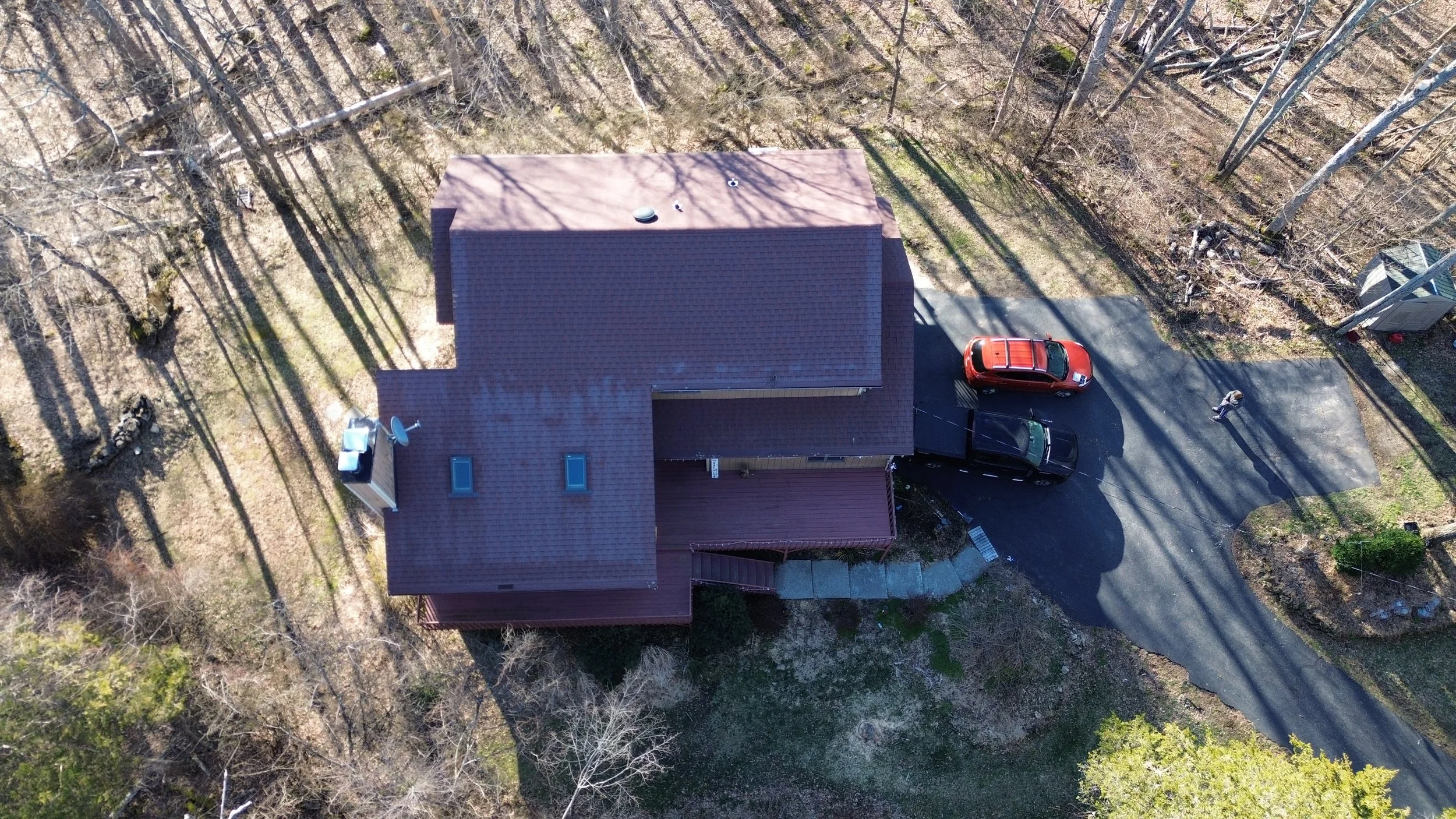 Aerial view of a house with a brown roof, driveway with two parked cars, and surrounding wooded area with leafless trees.