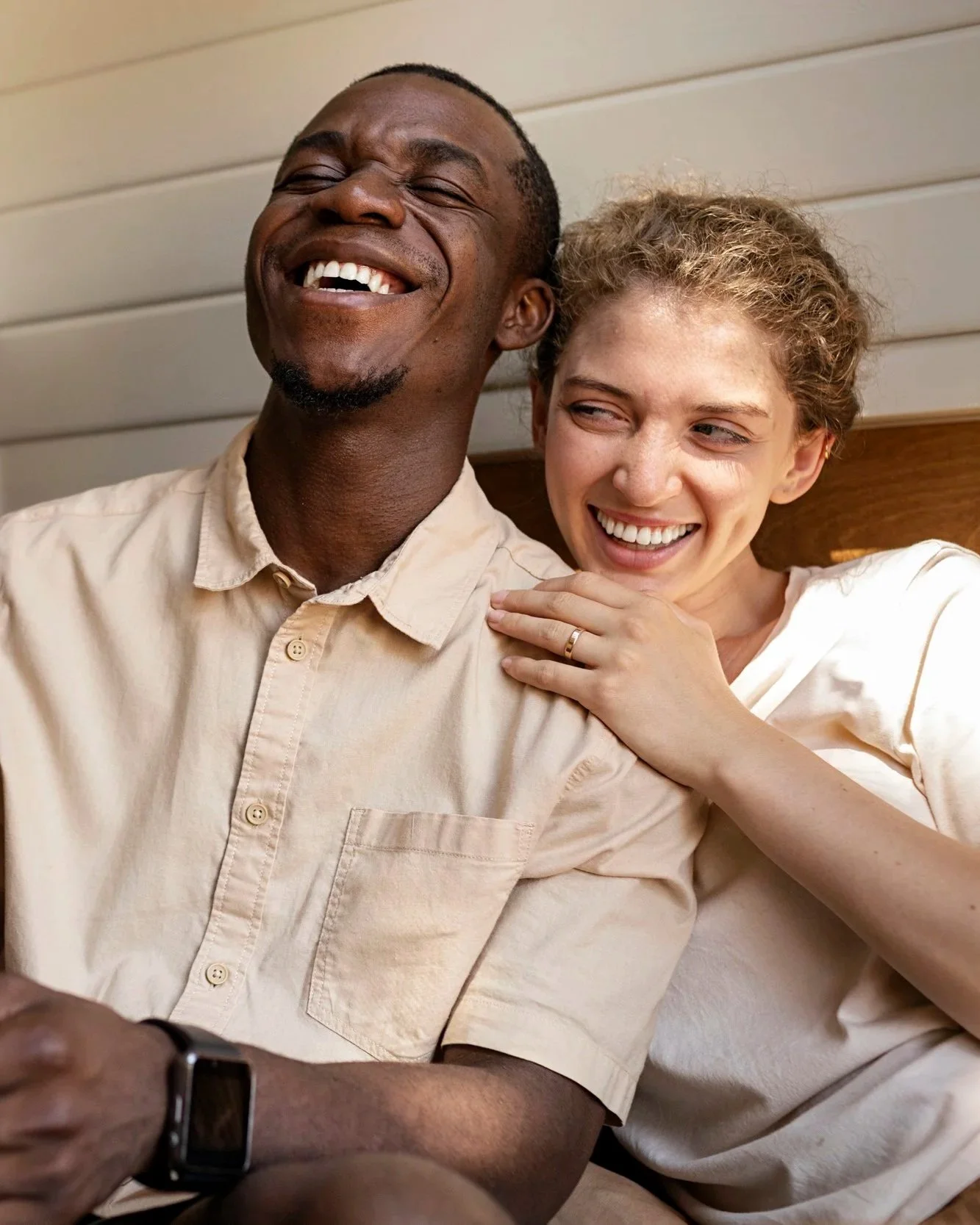 A man and woman sitting close together, laughing and smiling, with the woman gently touching the man's shoulder. The man is wearing a light-colored button-up shirt and a smartwatch, while the woman is wearing a light-colored top and has curly hair.