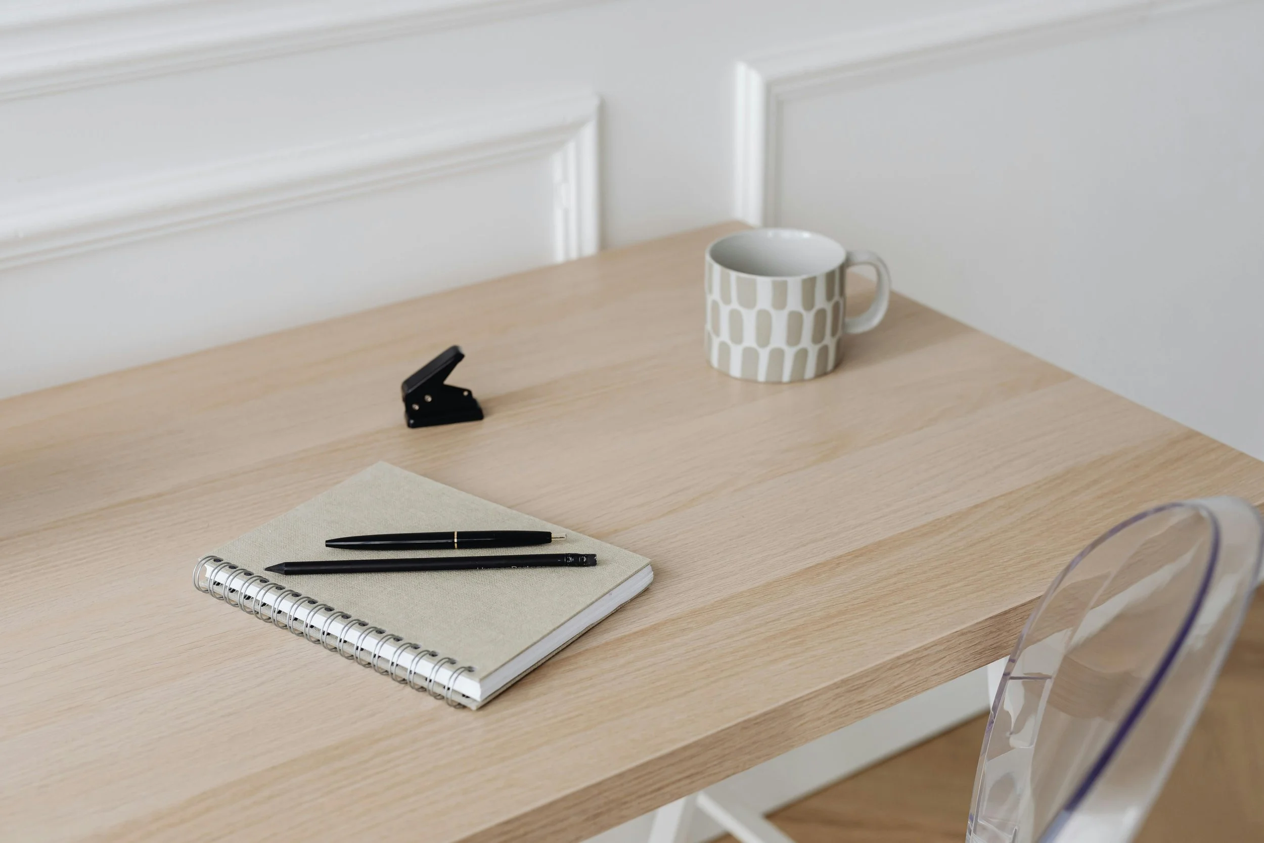 A wooden desk with a spiral notebook, two black pens, a white mug with a geometric pattern, a stapler, and a blank sheet of paper, against a background of a white wall with decorative molding.