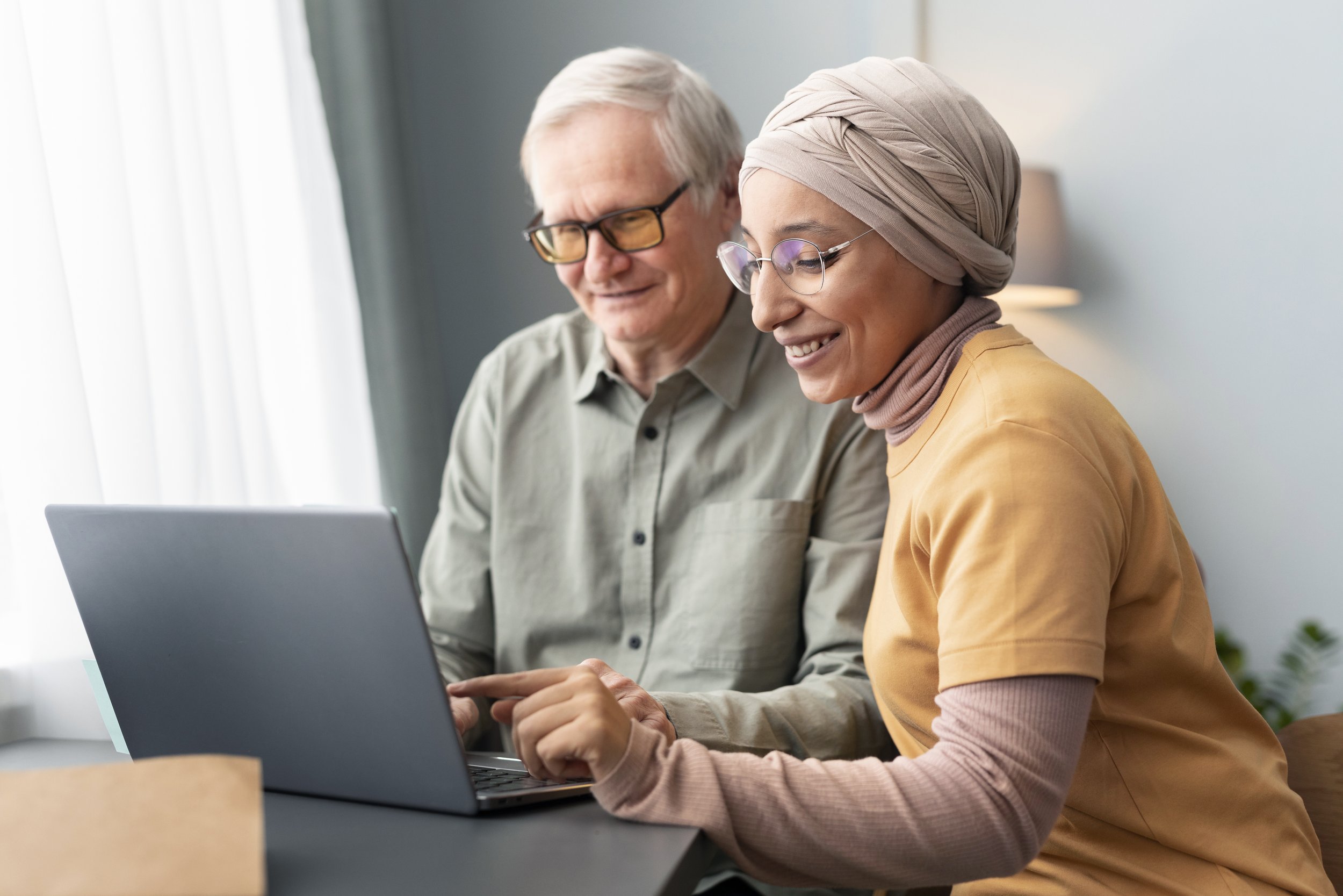 An elderly man wearing glasses and an elderly woman wearing glasses and a beige headscarf sitting together using a laptop in a bright room with curtains.