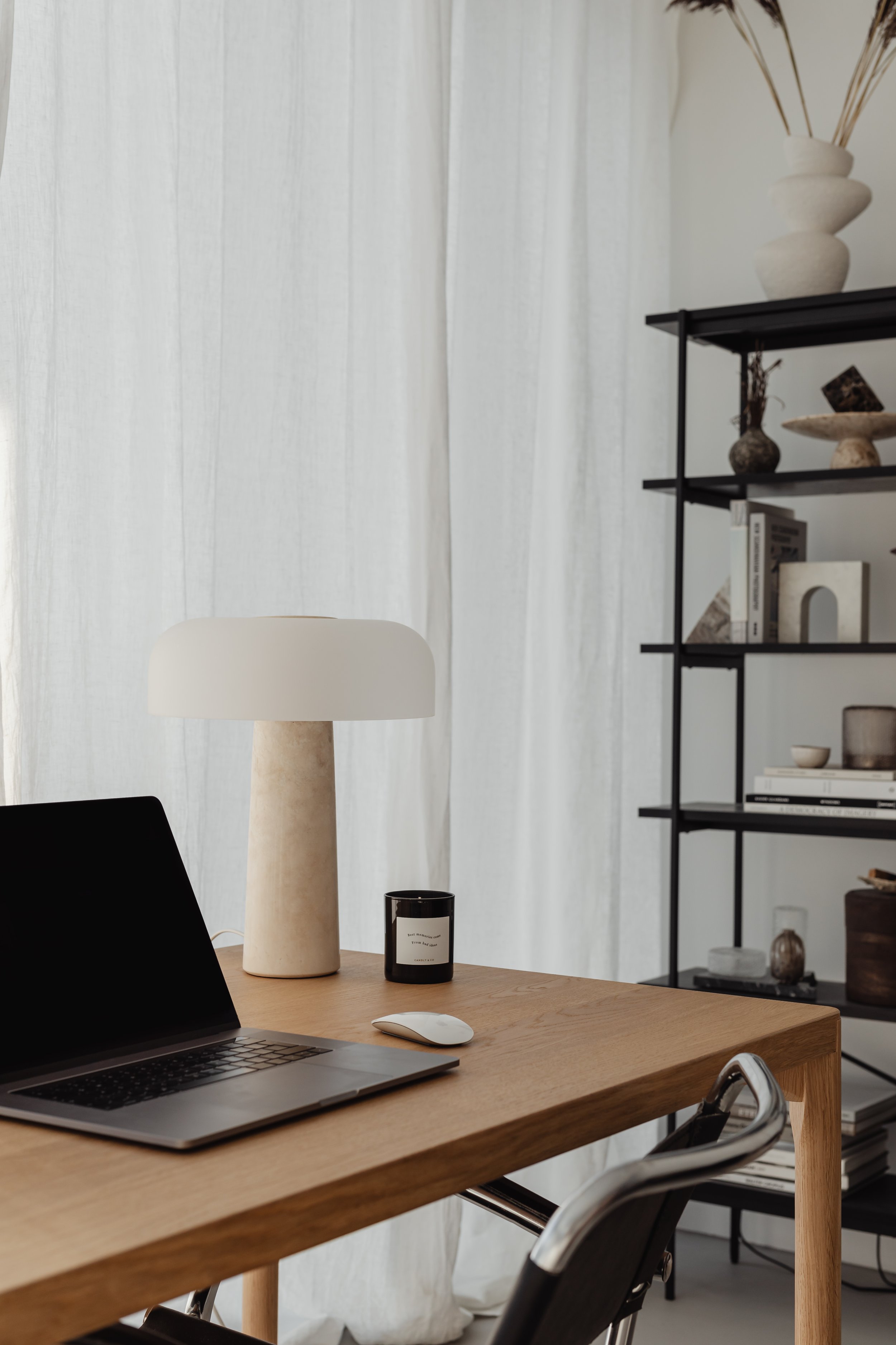A modern wooden desk with an open laptop, a wireless mouse, a black candle, a tall beige table lamp, and a black chair in a minimalistic office space. Behind the desk, there is a white curtain and a black metal shelf with decorative objects and books.