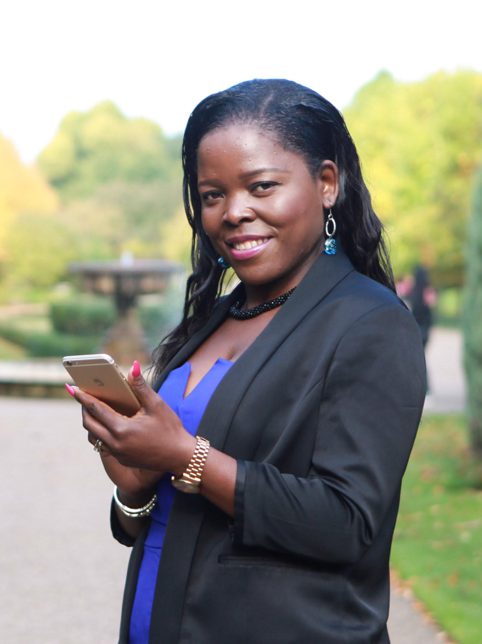 Smiling woman in professional attire holding a smartphone outdoors in a park.